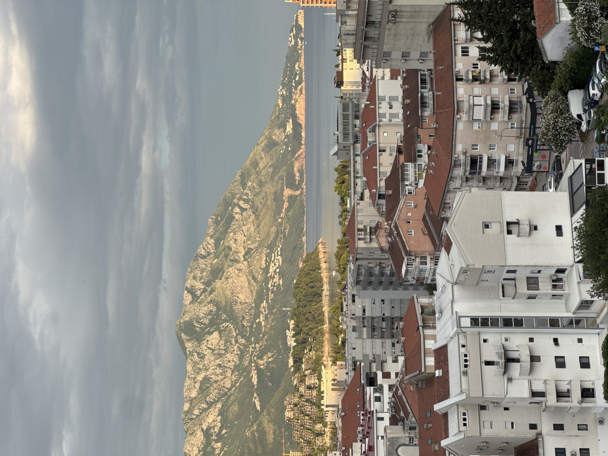 The image shows an elevated, slightly tilted view of Budva, Montenegro, facing inland from the coast. The scene is dominated by a large, mountainous terrain on the left side of the frame, transitioning to a thin strip of shoreline bordering the Adriatic Sea in the middle. Adjacent to the shoreline are dense rows of residential buildings, characterized by white facades and red tile roofs, tightly packed together. The buildings are densely interspersed with trees and foliage, particularly on the right side of the frame. The sky overhead is overcast, with cloud formations suggesting an impending change in weather. There are no individuals or other subjects visible within the frame.