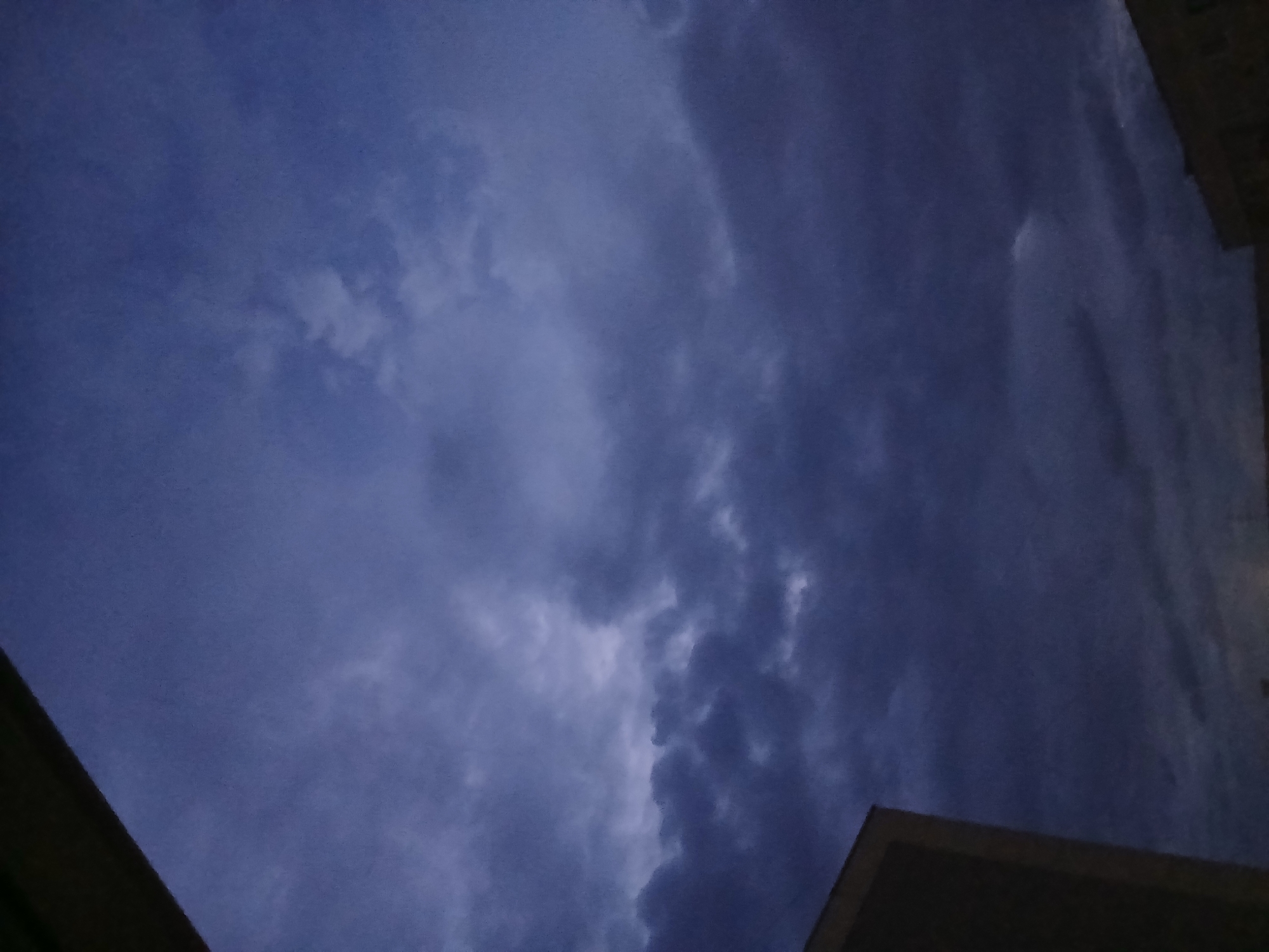 The image captures a cloudy sky over the city of Chaikovsky, Russia. The sky is predominantly a deep, dark blue, indicating either twilight or overcast conditions. Several layers of clouds are visible, with some appearing denser and darker, suggesting a buildup of storm clouds, while others are lighter and more diffused. In the lower left quadrant of the image, the corner of a building, possibly an apartment block, is visible. The building's facade appears to be a light color, contrasting with the dark sky. In the lower right, another building, also appearing to be of a residential nature, is partially visible. The scene is static, with no people or animals present. The overall atmosphere is somber and moody, characteristic of impending rain or a change in weather.