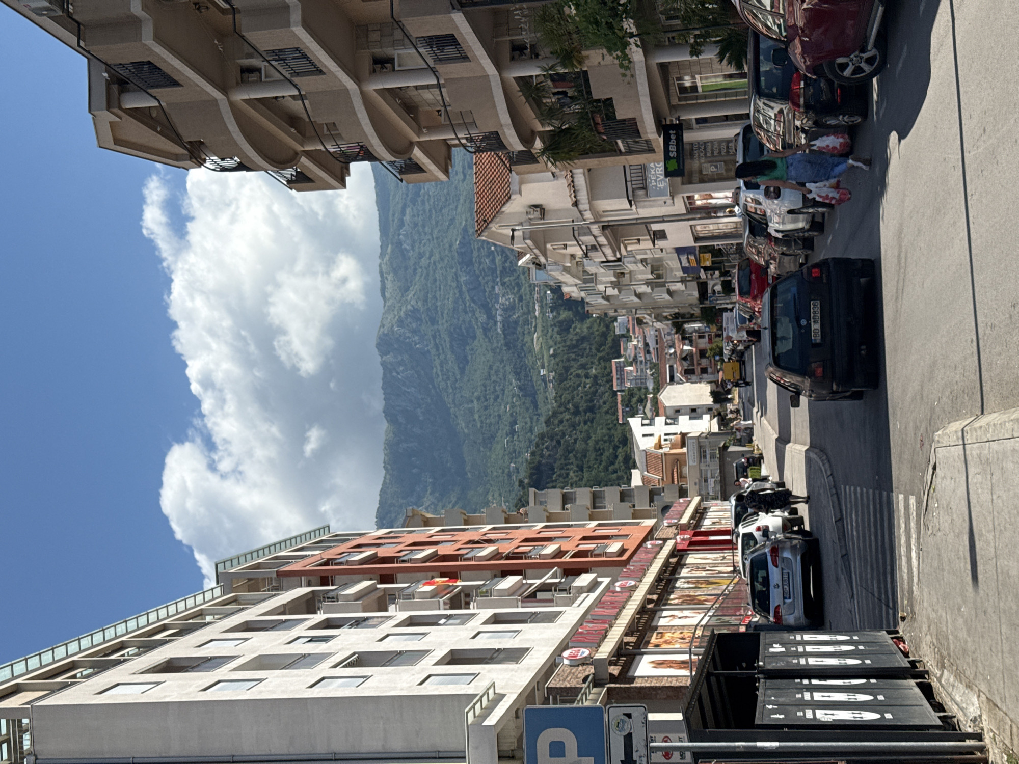 The image captures a vibrant, sunlit street scene in what appears to be an urban area with a dramatic mountainous backdrop, characteristic of Budva, Montenegro. The time of day is clearly daytime, evidenced by the bright blue sky adorned with fluffy white clouds and strong, distinct shadows cast by buildings and vehicles, indicating sunny weather.
In the foreground, a paved street runs along the bottom, lined with numerous parked cars. Among these, a dark grey car with the license plate "BO AD836" is prominently featured in the lower right, and further down the street, a silver car with the license plate "BO 16311" is visible. A pedestrian is seen walking on the sidewalk between parked cars, carrying bags. Along the street, there are large, dark grey trash or recycling containers.
The left side of the street is flanked by multi-story buildings of varying architectural styles. Some are modern, featuring light-colored facades and glass balconies, while others have more traditional designs with reddish-brown tiled roofs. The lower levels of these buildings house commercial establishments, with visible signage. Text includes "SBbet" and "PEKA EVRC" on one building, and a blue "P" sign, likely indicating parking, is visible near the bottom left.
Dominating the background are majestic, verdant mountains covered in dense forestation, stretching across the landscape under the bright sky. The scene presents a lively urban environment set against a striking natural landscape.