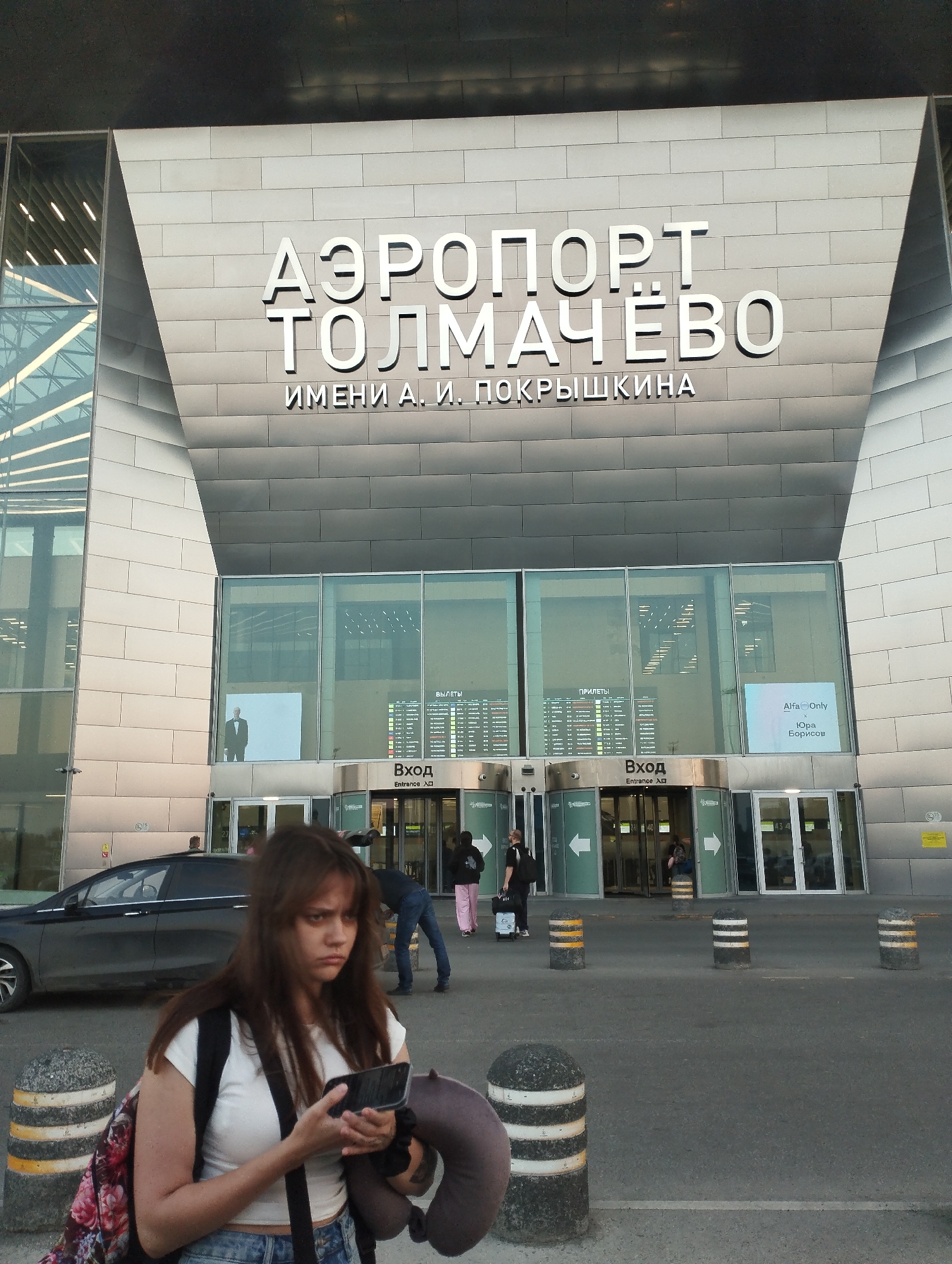 The image shows the Tolmachevo Airport in Ob, Russia, with a young woman in the foreground holding a smartphone and looking down. She is wearing a white t-shirt, blue jeans, and a backpack with a floral pattern. A neck pillow is slung over her shoulder. In the background, the modern facade of the airport building is visible, featuring large glass windows and metallic cladding. Signage on the building reads "AEROPORT TOLMACHEVO" and "IMENI A.I. POKRYSHKINA," indicating it is Tolmachevo Airport named after A.I. Pokryshkin. Several revolving doors are visible, with people entering and exiting. A black car is parked to the left of the entrance. Bollards with yellow and white stripes are scattered on the pavement. The sky is visible at the top, with a shadow cast by the building. The overall scene depicts a typical airport environment with travelers.