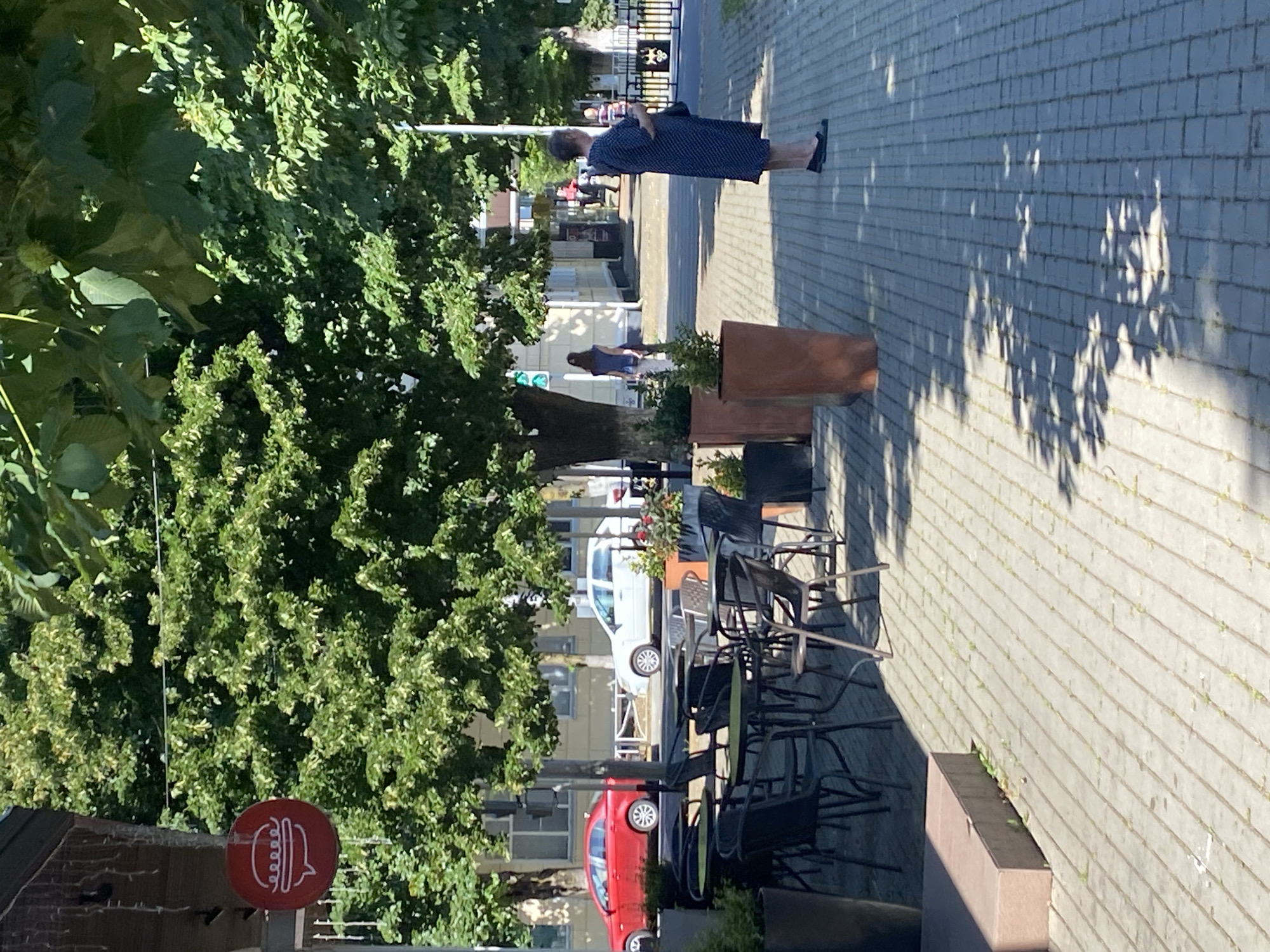 The image depicts an outdoor seating area for a restaurant, likely located in Maykop, Russia, given the context. In the foreground, several black metal tables and chairs are arranged on a paved stone patio. Large planters with greenery are visible, along with a red circular sign with a white hamburger icon, suggesting a fast-food establishment. Beyond the patio, a street is lined with trees, and a red car and a white car are parked or driving on the road. In the mid-ground, several people are present. A woman in a dark blue patterned dress stands on the sidewalk, facing away from the camera. Further in the background, other individuals can be seen walking. The environment appears to be a sunny day, with shadows cast by the trees and awnings across the patio.