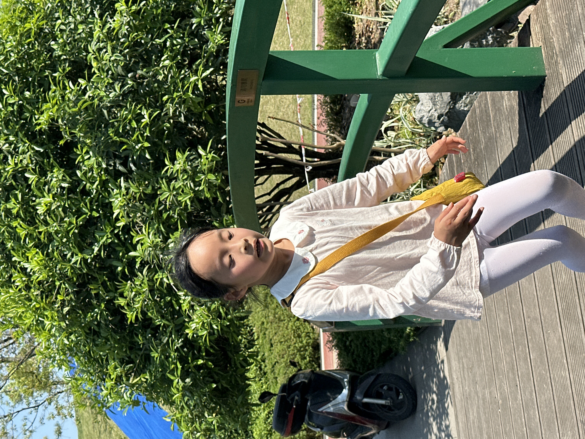 The image shows a young Asian girl sitting on a green wooden structure, possibly a bridge or elevated walkway, in what appears to be a park or garden setting. She is wearing a light-colored, long-sleeved shirt and white leggings, with a yellow bag slung across her body. Her eyes are partially closed, and she is reaching out with her right hand.
Behind the girl, there is dense green foliage, and a glimpse of blue sky and what appears to be a blue structure or tarp. A scooter is parked near the base of the structure on the right.
Based on the prompt, this image was taken in the city of Jiangjin, China.
