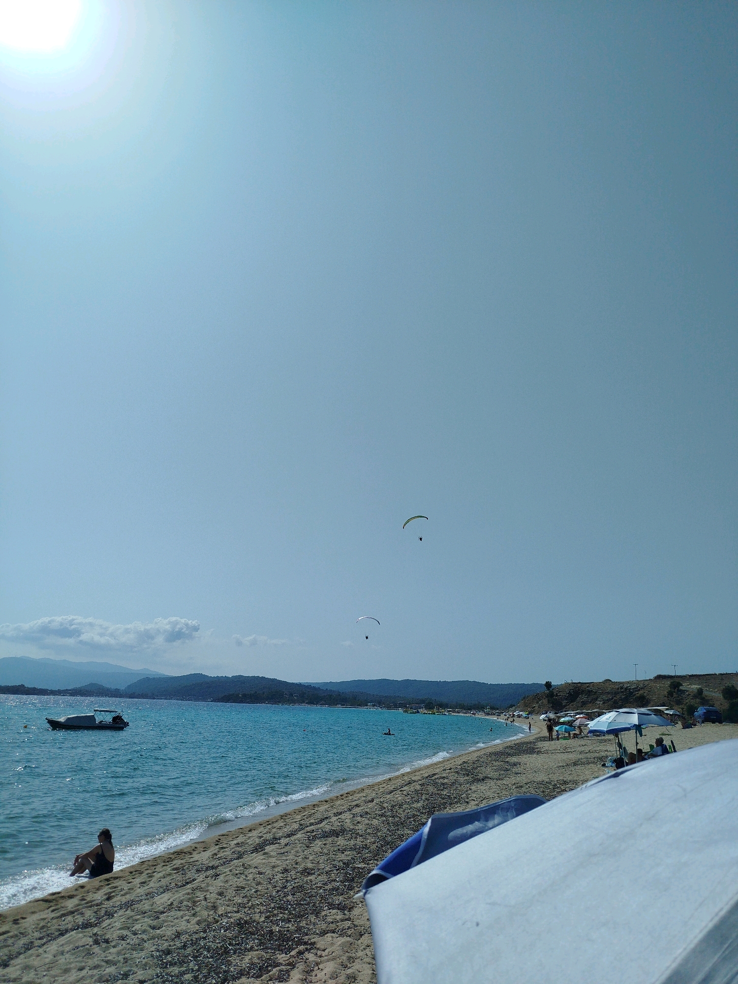 The image depicts a sunny day at a beach in Sithonia Municipal Unit, Greece. The clear turquoise water of the Aegean Sea gently laps the sandy shore. A person is seated on the sand at the water's edge, facing the sea. In the distance, a small boat with a canopy is anchored in the water. The beach is dotted with umbrellas, some occupied by people, indicating a leisurely beach scene. Further down the coastline, a paraglider is visible in the sky, enjoying the aerial view. The background features rolling hills covered in lush green vegetation. The overall atmosphere is one of relaxation and summer recreation.