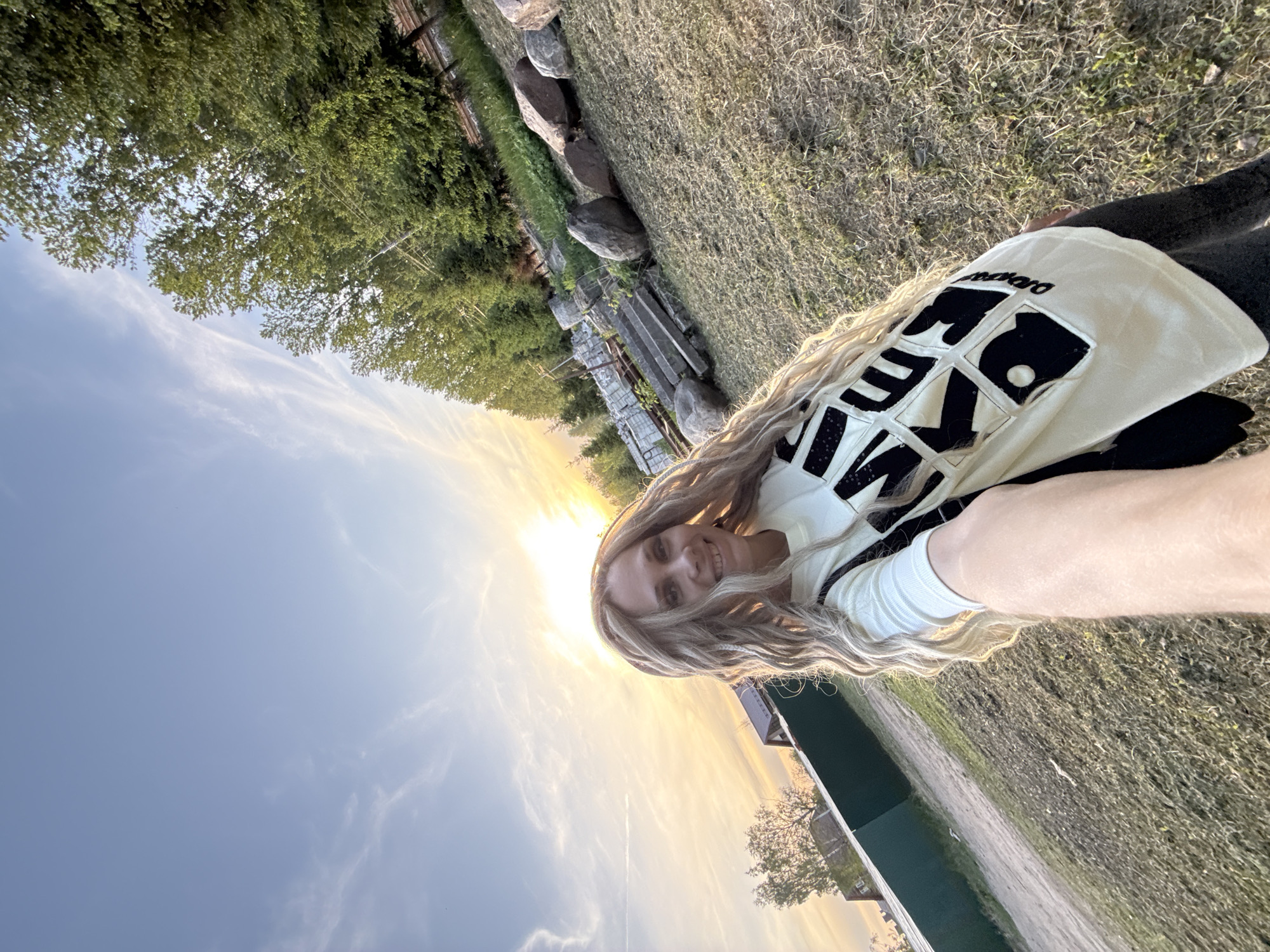 A medium shot captures a blonde woman smiling directly at the camera. She is wearing a light-colored shirt with dark, possibly black, appliqués or lettering on the front. The background is split between a clear, blue sky at the lower left and a bright, golden sunset. A line of trees is visible at the upper left, and a grassy field occupies the upper right. In the middle ground, there is a line of stones, and a path or road runs through the lower right portion of the image. The location appears to be in the city of Rusanovo, Russia.