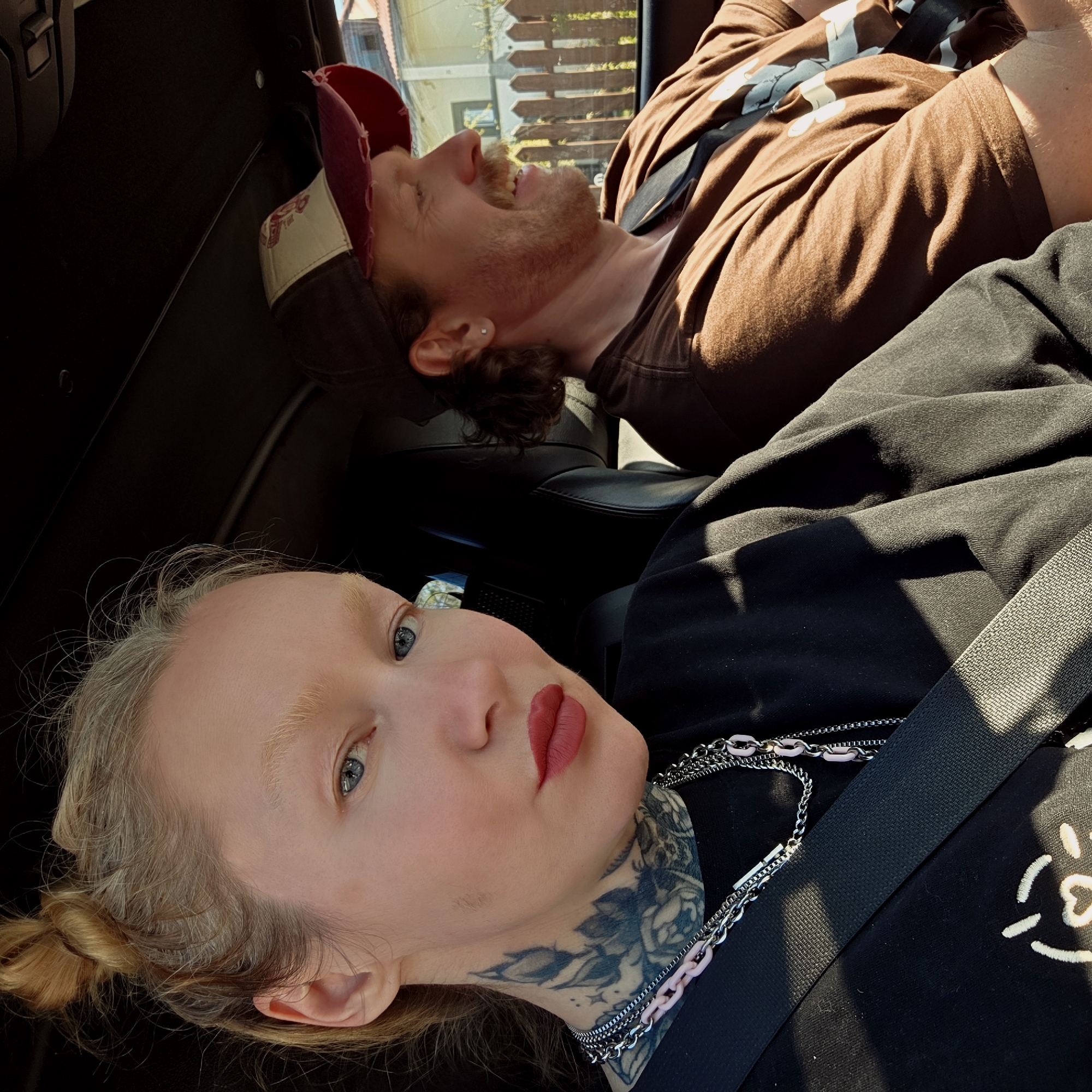 Two people are in the front seats of a car. The person in the foreground is fair-skinned, with blonde hair tied in a bun, blue eyes, and red lipstick. They have tattoos on their neck and are wearing a black shirt with a white design. The person in the background has red hair, a baseball cap, and is wearing a brown shirt. Both are wearing seatbelts. The location appears to be Камянюцкі сельскі Савет, Belarus, based on the view through the car window showing a fence and foliage typical of a rural or suburban area.