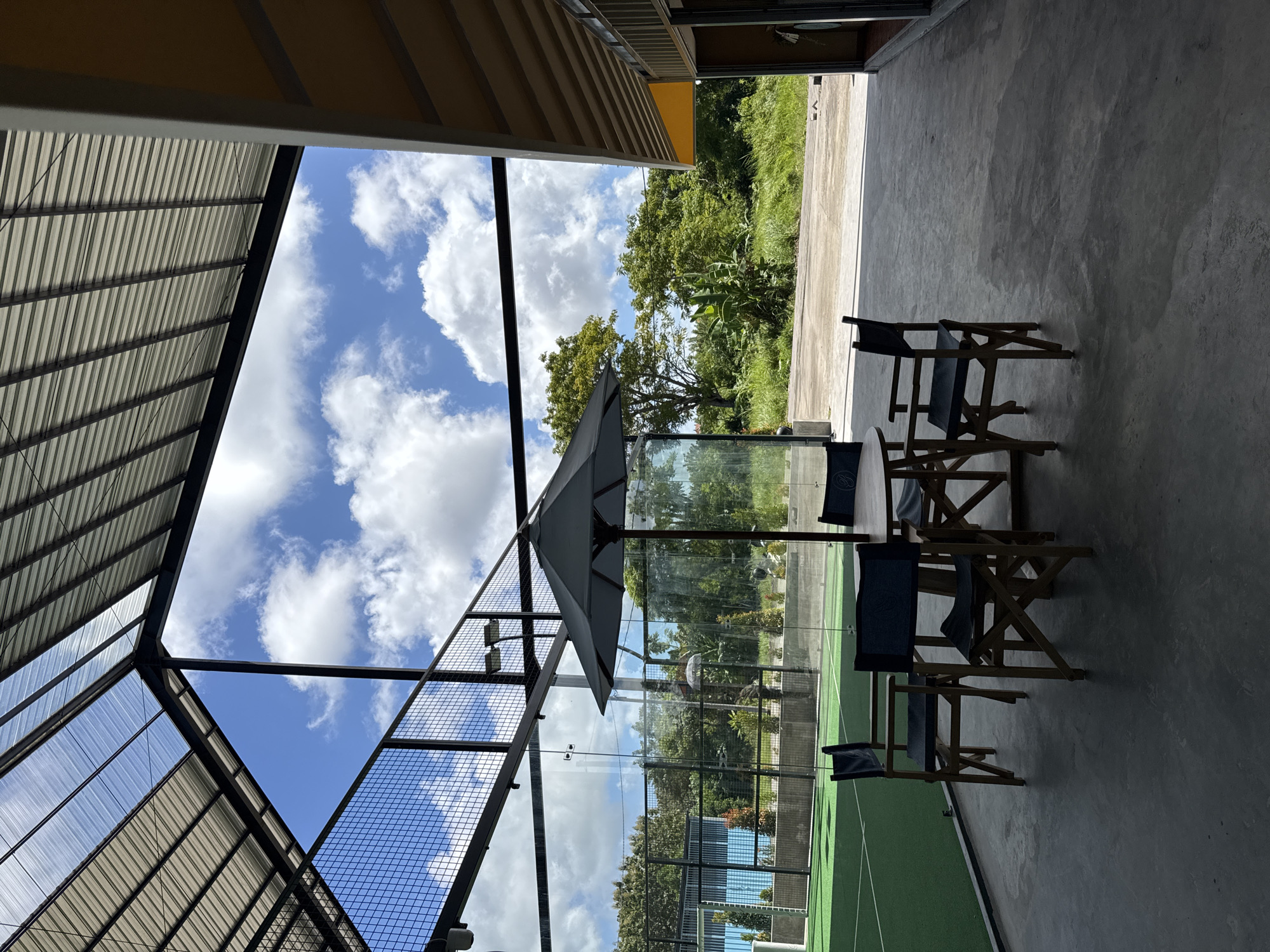 A partially enclosed outdoor seating area is situated in Unknown, Unknown, as indicated by the presence of a padel tennis court. The area features four wooden folding chairs with dark blue seat cushions arranged around a light-colored umbrella shading a table. The seating area is situated on a gray concrete surface, part of which is under a structure with a corrugated metal roof. The area overlooks a padel tennis court enclosed by a wire mesh frame and glass panels, reflecting the cloudy sky. Beyond the court, trees and greenery suggest a park or undeveloped area. The seating area is open on one side, revealing a concrete path leading to an open area with vegetation.