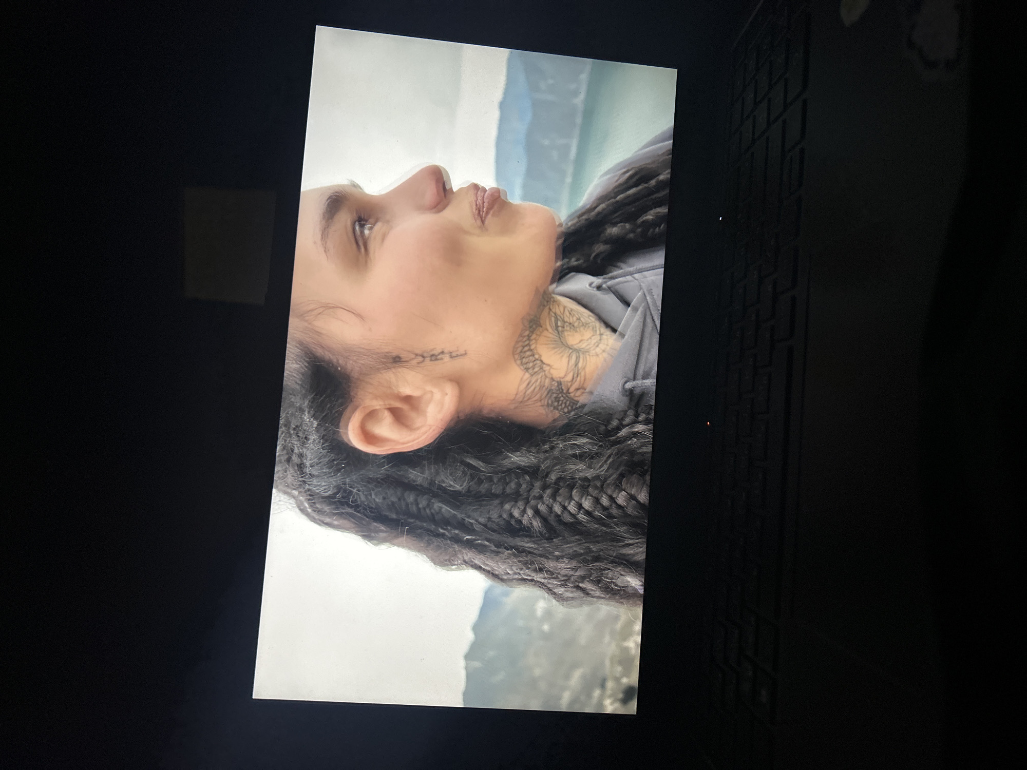A digital photograph shows a woman's profile against a blurred background of water and hills, possibly a coastal or lakeside scene. She has dark hair styled in braids or locs, and tattoos are visible on her neck and near her ear. She wears a grey hooded sweatshirt. The photo appears to be taken of a screen, with part of a black keyboard visible on the right. The location, Kalchevo, Bulgaria is not evident from the visual information in the image.
