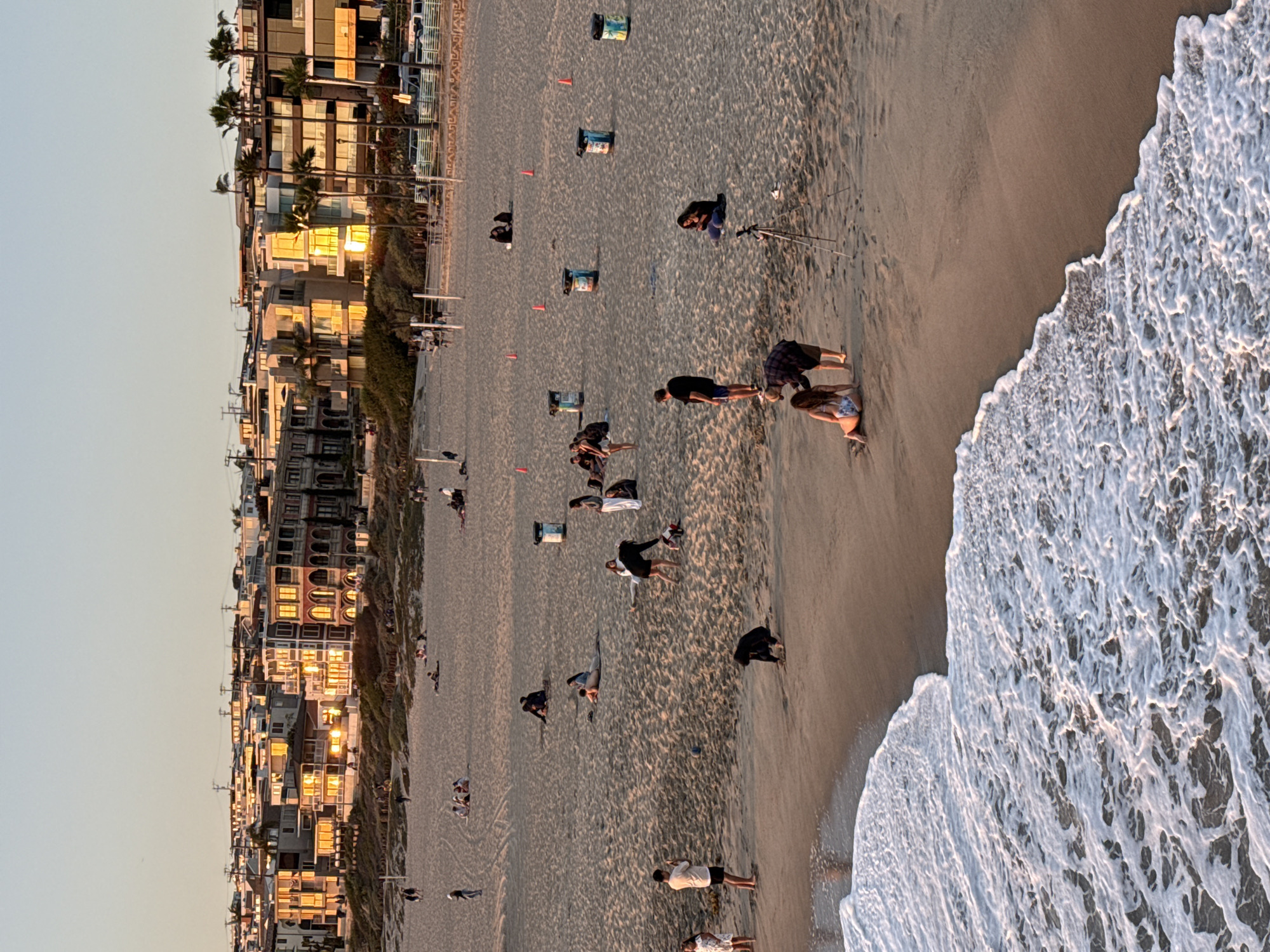 This is a medium-angle, full shot of a beach in Manhattan Beach, California, during sunset. The warm light of the setting sun illuminates the buildings in the background, casting long shadows across the sand. The beach is populated with people engaged in various activities. Some are walking, some are sitting and relaxing, and others appear to be posing for photographs. The ocean is visible in the foreground, with gentle waves lapping at the shore. The scene evokes a sense of calm and leisure, typical of a beachside community at the end of the day.