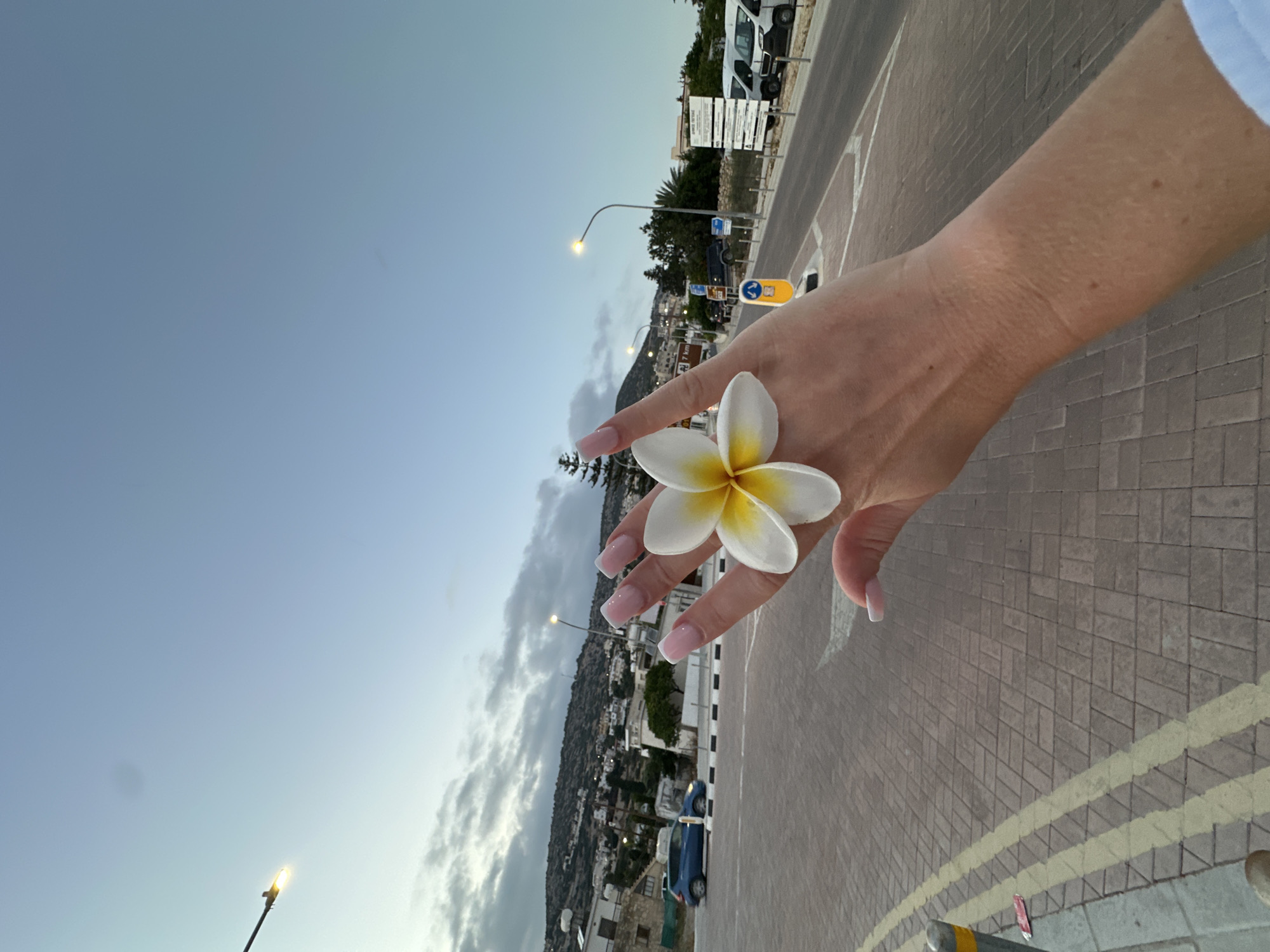 The image shows a close-up of a person's hand with a white and yellow plumeria flower resting on their fingers. The person has long, light pink French-tip acrylic nails. The hand is positioned in the foreground against a backdrop of a street scene. In the background, there is a paved road with yellow double lines, a sidewalk, streetlights, parked vehicles including vans, and buildings on a hillside. The sky is a pale blue with scattered clouds, suggesting it is either early morning or late evening. The location is identified as the city of Peyia, Cyprus.