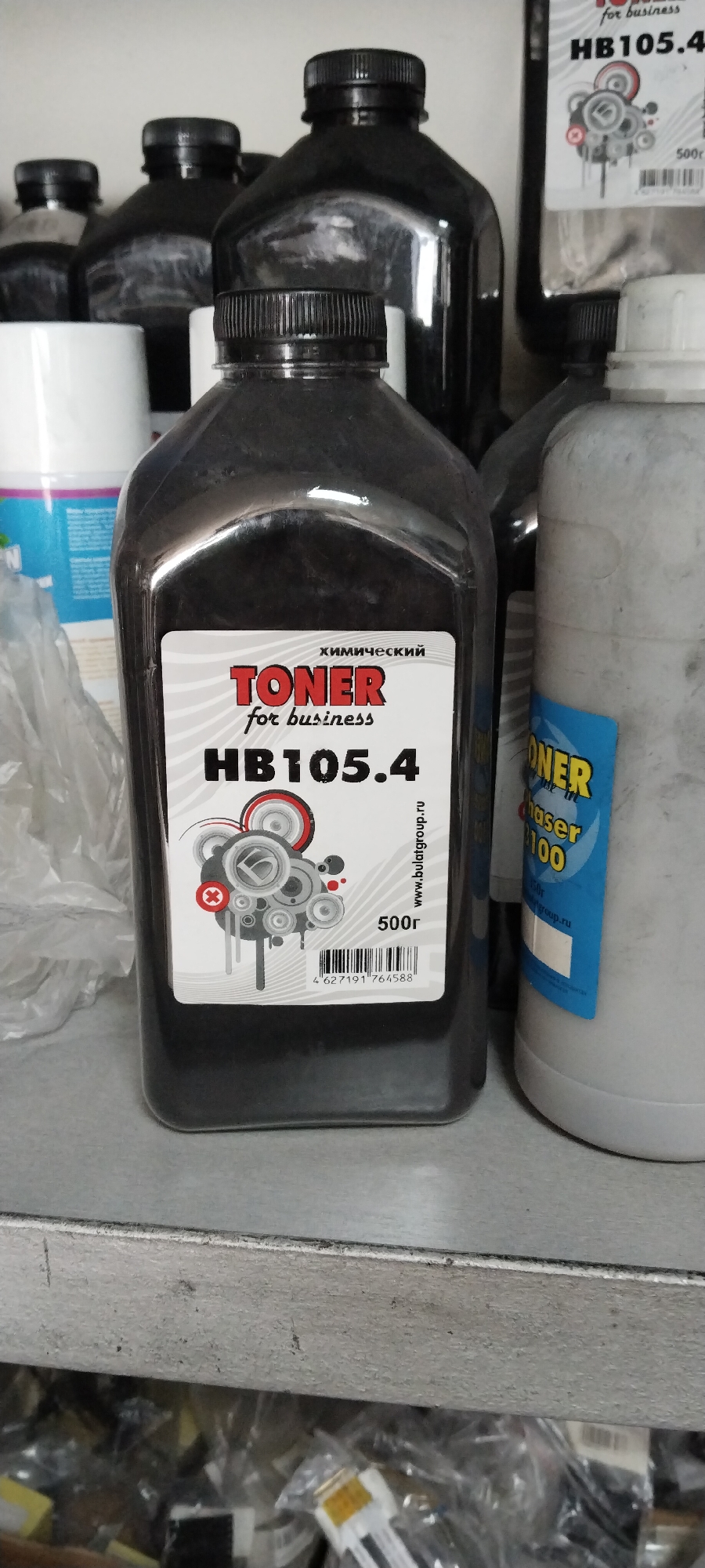A close-up, eye-level, interior shot captures multiple black plastic bottles filled with toner powder, likely on a shelf in a commercial or storage environment. The primary focus is a bottle in the center foreground, displaying a white label with "TONER for business HB105.4" prominently printed in red and black. To the right of this bottle is another, lighter-colored bottle with a blue and yellow label that reads "TONER chaser 100". The background is out of focus, showing additional black bottles of varying sizes and shapes, along with a white cylindrical object on the far left. The lighting appears to be artificial, casting soft shadows. The shelf itself is a weathered, metallic surface. The scene suggests a retail setting, warehouse, or office supply storage area. The location is not specified.