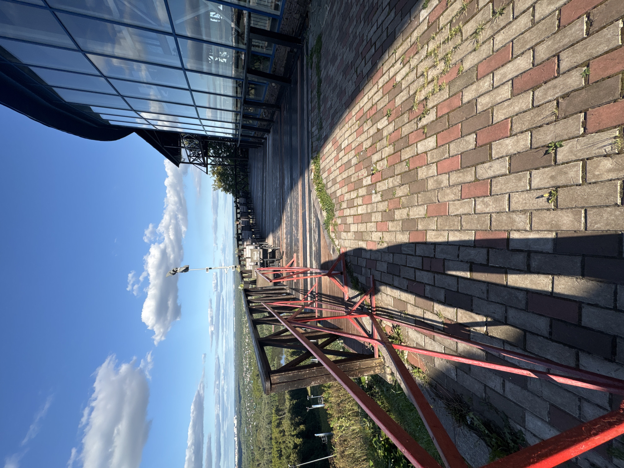 The image depicts an outdoor seating area with a paved walkway and wooden decking, overlooking a lush green landscape with a river and a distant city. The architecture of the building suggests a modern establishment with a curved facade of large glass windows. The scene appears to be set in the city of Ufa, Russia, possibly at a viewpoint or restaurant on a hill. The sky is clear blue with scattered white clouds, indicating fair weather. There are no people visible in the image, and the area seems quiet and serene. The red metal railing in the foreground suggests a safety barrier along the edge of the elevated area.