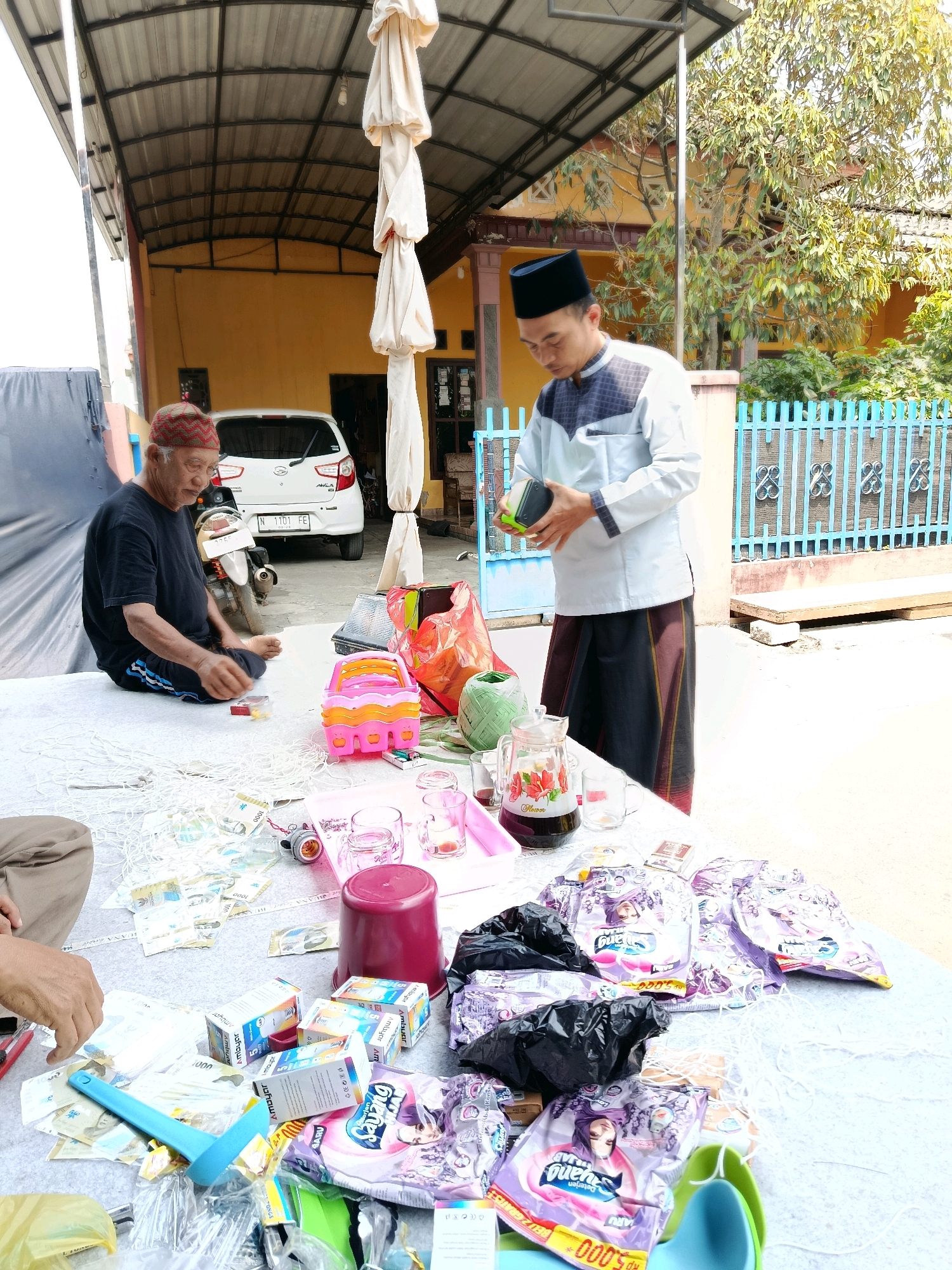 In Bululawang, Indonesia, an outdoor scene unfolds under a metal roof structure. Two men are present. The first, an older man with graying hair and glasses, sits on a white cloth-covered surface, wearing a black t-shirt and a red and white patterned beanie. He is looking down, with his hands near a collection of what appear to be Indonesian Rupiah banknotes and some small items.
The second man, younger and wearing a black peci hat, a light-colored patterned shirt, and a dark sarong, stands to the right. He is holding a black and green object in his hands.
A white car is parked behind the older man. The ground is paved. In the foreground, a large white cloth covers the ground, on which various items are spread out. These include numerous Indonesian Rupiah banknotes, several packs of cigarettes, some small boxes labeled "Ambujur," a pink plastic toy, a large red plastic container, a pitcher with dark liquid, glasses, a green woven basket, and several purple-colored packages with images of women. A blue plastic scoop and other assorted plastic items are also visible. A blue metal fence is in the background, with foliage and a yellow building beyond it. The environment suggests a gathering or event.