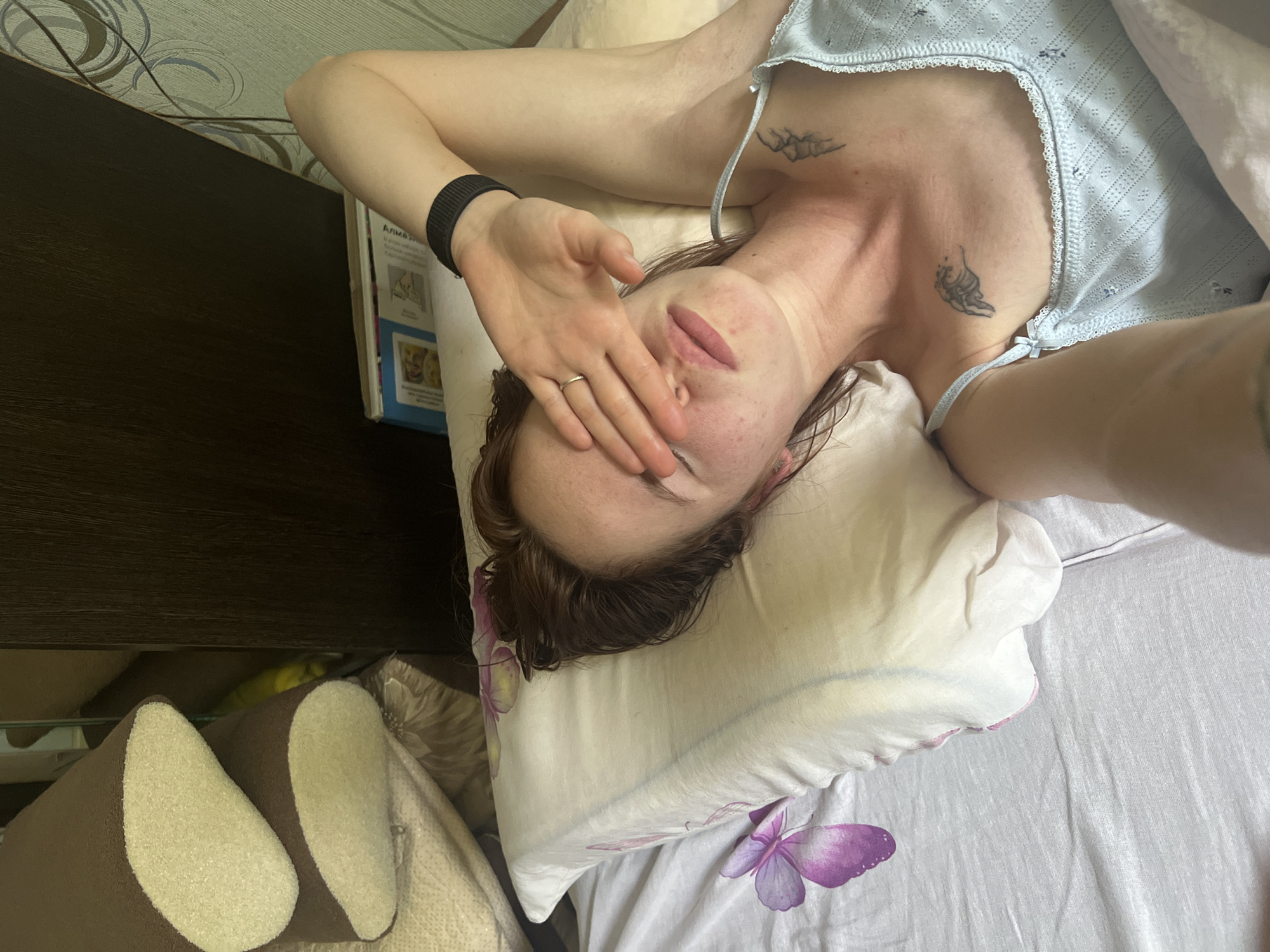 The image shows a woman lying in bed, covering her eyes with her hand. She is wearing a light blue camisole. There are tattoos on her chest and neck. The bed has a white pillow with a purple flower design and a white sheet. In the background, there is a dark brown wooden furniture piece and a wall with a patterned wallpaper. The scene appears to be indoors, possibly a bedroom. The image is a selfie, taken from a high angle.