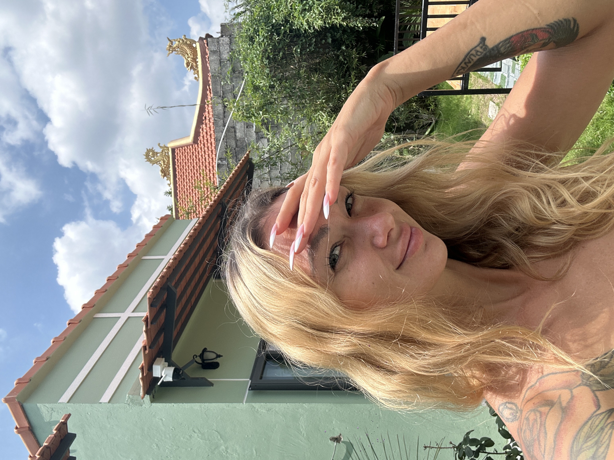 A close-up selfie captures a fair-skinned woman with long, wavy blonde hair and light green eyes, shielding her face from the sun with her right hand. She is wearing a strapless top, revealing tattoos on her left shoulder and right arm. The background shows a light green building with a terracotta-tiled roof and a grey stone wall with a lush green bamboo garden. The sky is bright blue with scattered white clouds. The scene is likely set in Mũi Né, Vietnam, given the architectural style and vegetation.