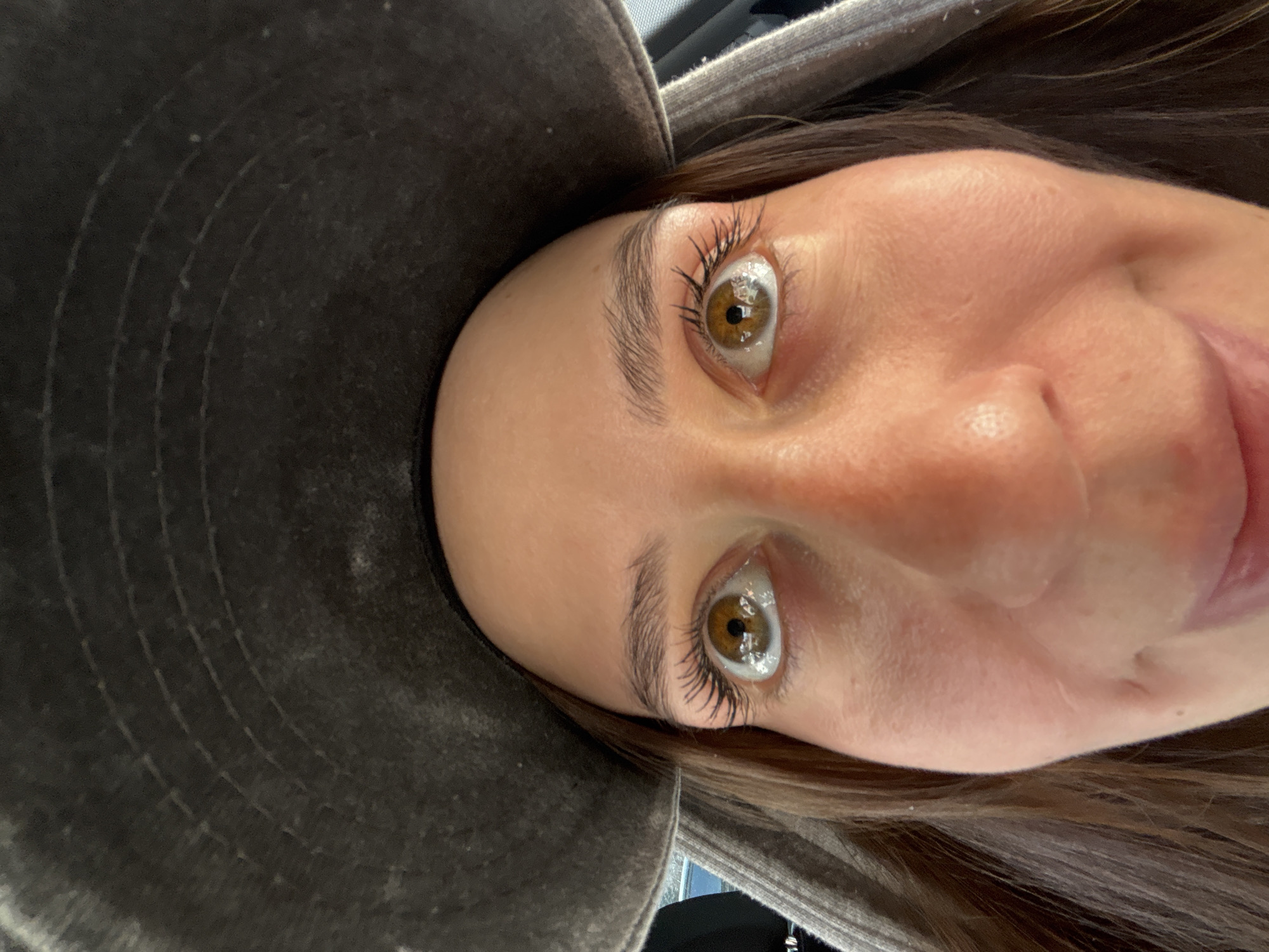 The image displays a close-up, low-angle portrait of a person, presented in a rotated orientation such that their head is tilted downwards towards the left. The subject, appearing to be female, looks directly into the camera with wide, light brown eyes. Reflections of multiple light sources and what appears to be a window or car interior are prominently visible in their eyes.
The person has long, dark eyelashes, likely enhanced with mascara, and well-defined, dark eyebrows. A subtle, gentle smile is visible on the right side of their mouth. Dark brown hair cascades down the right side of the frame. They are wearing a dark, textured, possibly distressed fabric hat or hood, which covers the upper left portion of their head and frames their face.
The background is indistinct, consisting mainly of the dark fabric of the hat/hood and hints of a dark, blurry setting. There are no specific location cues visible within the image itself to confirm the provided context of Krasnoyarsk, Russia. The time of day and weather conditions are not discernible. No text is visible in the media file.