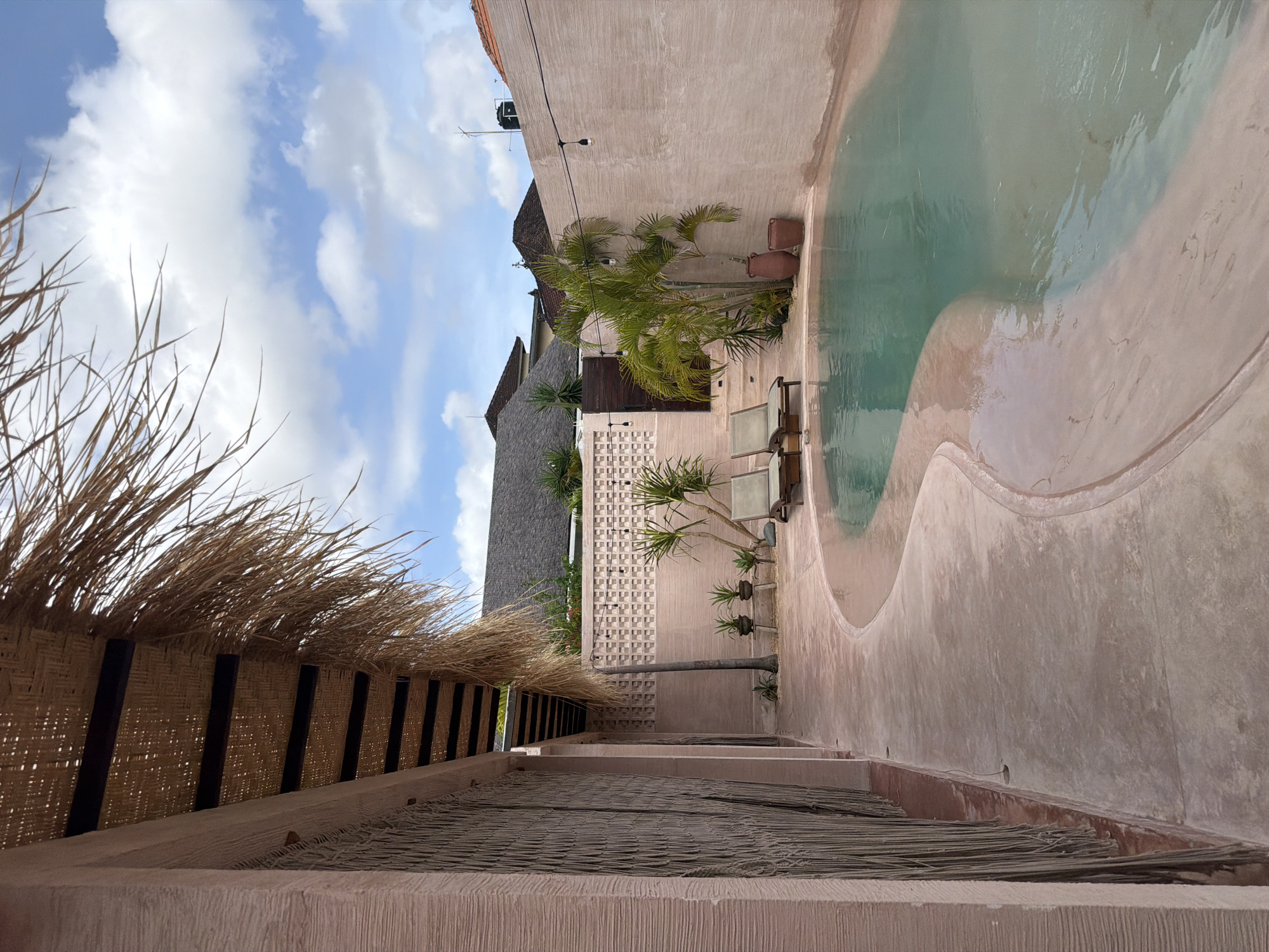 The image depicts a hotel in North Kuta, Indonesia, featuring a unique architectural design. The foreground showcases a woven thatched roof structure supported by dark-colored beams, indicating a traditional or rustic design element. Beyond this, there is a walkway leading towards a swimming pool with a freeform shape, characteristic of modern resort designs. The pool is surrounded by light-colored textured walls, with some vegetation, including palm trees and potted plants, providing a touch of greenery. The architectural style of the buildings in the background shows a mix of modern and traditional elements. The sky is visible above, displaying scattered clouds and blue patches. Overall, the image conveys a sense of a tropical resort with a blend of traditional and modern design elements.