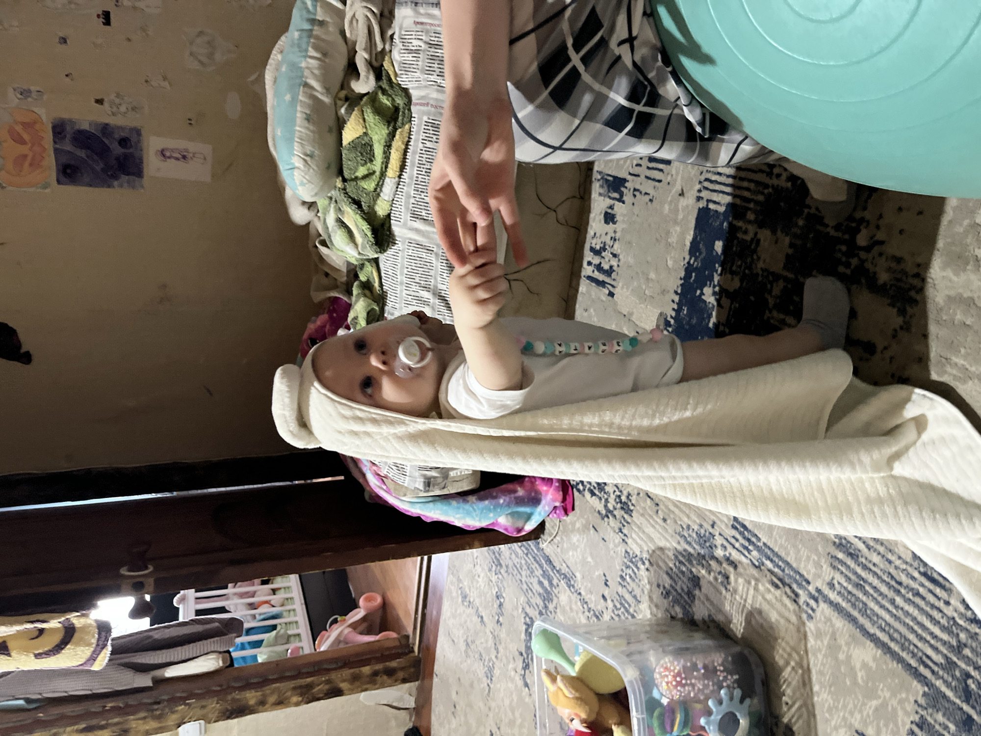 A baby lies swaddled on a patterned rug. An adult's hand is holding the baby's hand. A green exercise ball is nearby. The location is Vesyoloye, Russia.