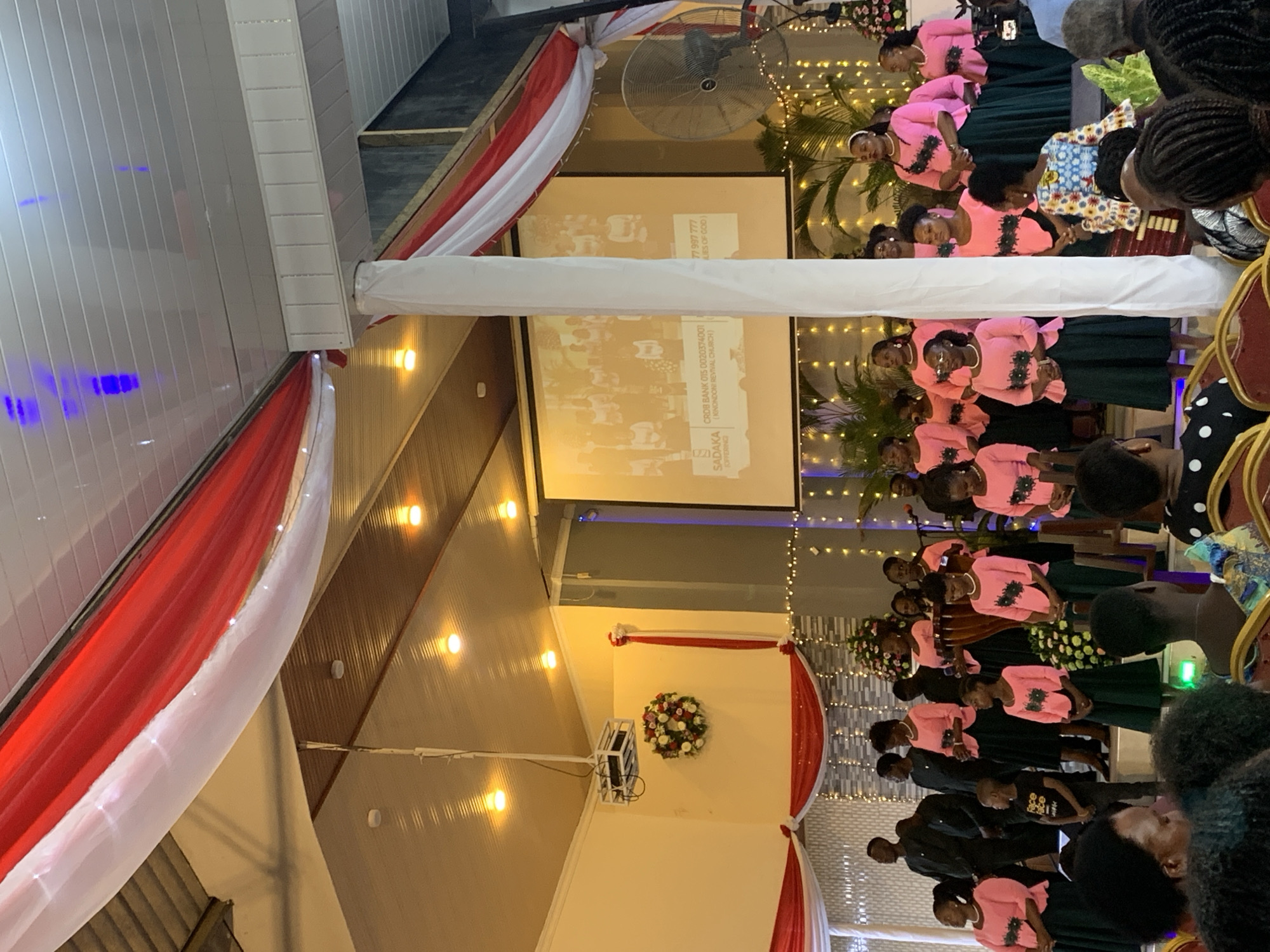 The image shows a group of people, primarily women, gathered indoors, likely in a church setting. The majority are wearing pink tops and dark green skirts, possibly a choir or performance group. There is a screen displaying information including the names "CROB BANK" and "SADAKA (KINONDONI REVIVAL CHURCH)." Based on the name, and the fact that it says "Offering" on the screen, it is possible that this is a location in Kinondoni, which is a district in the city of Dar es-Salaam, Tanzania. The environment is decorated with red and white fabric, plants, and string lights. A large fan is visible in the background. The audience is partially visible in the foreground.