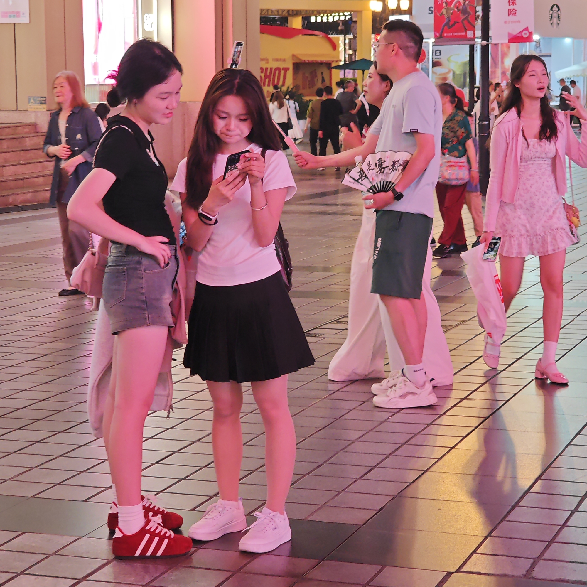The photograph depicts an outdoor scene in the urban environment of Yuzhong District, China. Two young women stand close together, one dressed in denim shorts, a black t-shirt, and red Adidas sneakers, and the other in a white t-shirt, black pleated skirt, and white sneakers; the second woman is looking at a smartphone. A man stands to their right, holding a folding fan and wearing a light blue t-shirt and dark green shorts; another woman stands behind him. In the background, additional pedestrians can be seen walking along the tiled pavement. A building with signage in Chinese characters is visible in the upper part of the frame, along with colorful banners and advertisements. The overall color scheme of the photo appears to be pink.