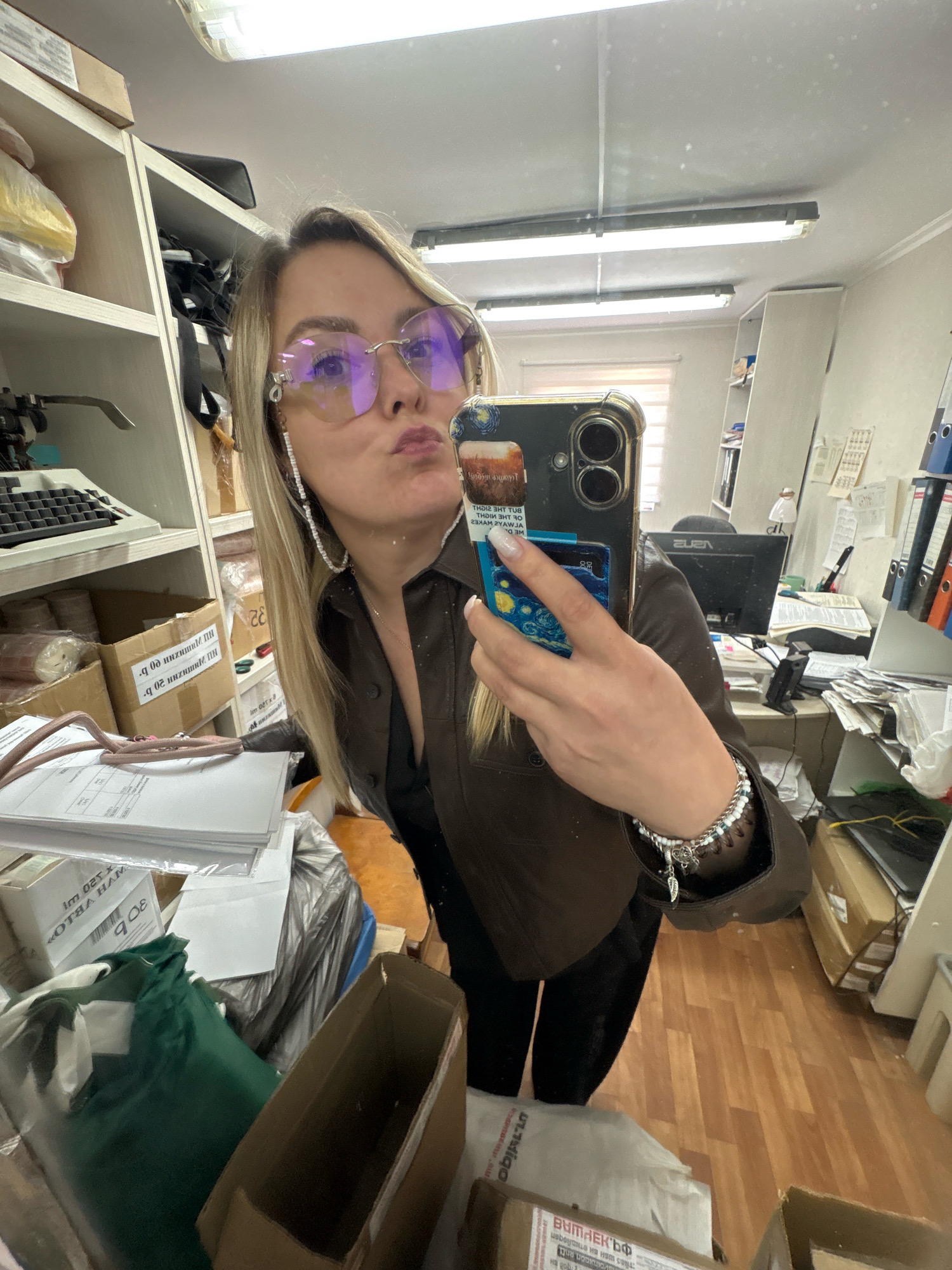 The image shows a woman taking a mirror selfie in what appears to be an office or storage room. She is wearing a brown jacket, black pants, and large, light purple tinted glasses. She has blonde hair and is making a kissing face. She is wearing a beaded bracelet on her left wrist and hanging earrings. Her phone, with a "Starry Night" case and other stickers, is visible as she holds it up to take the photo. The background is cluttered with shelves filled with boxes, papers, and office supplies. There is also a desk with a computer monitor and various items. The room appears to be indoors, with fluorescent lighting on the ceiling. The floor is made of wooden planks. There is no information in the image to suggest the location is in Noviy, Russia.