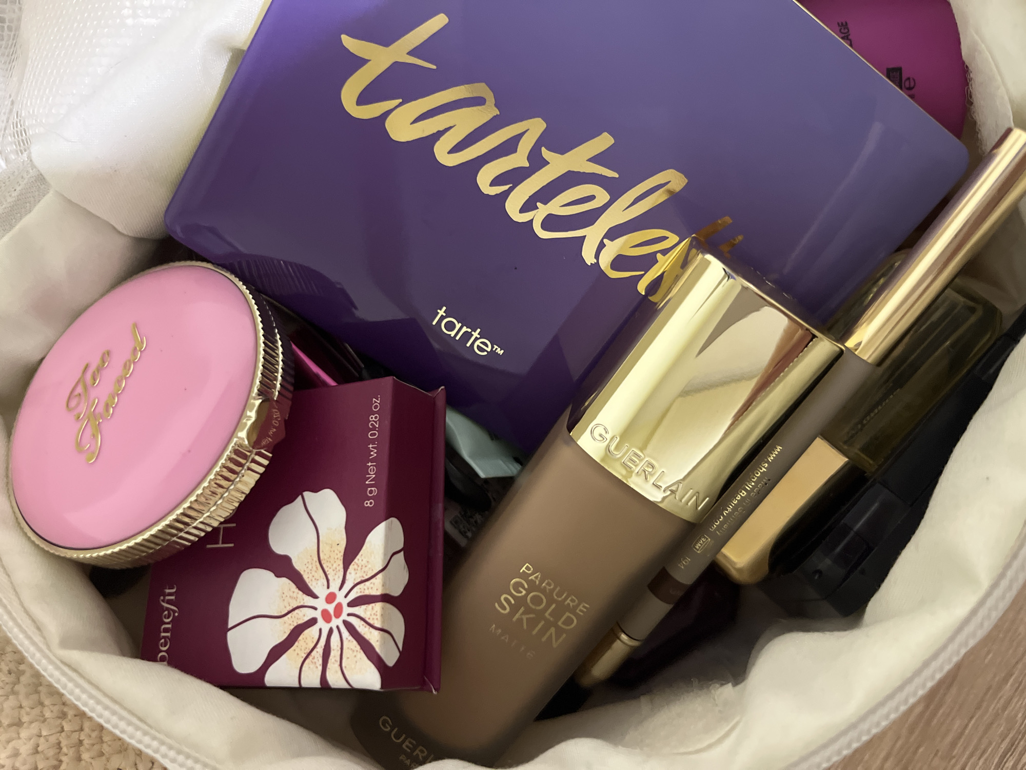 A high-angle, close-up shot showcases an assortment of makeup products nestled within a white zippered makeup bag. In the foreground, a gold-capped tube, identified as "PARURE GOLD SKIN" by GUERLAIN, lies horizontally. Partially visible beneath it are a dark-tipped eyebrow pencil and a black mascara tube. Behind these, a rectangular purple eyeshadow palette with gold lettering spelling out "tarte" is angled prominently. To the left of the palette, a small rectangular box with a floral design and "benefit" branding is visible. Topping the collection is a round, pink compact with gold "Too Faced" lettering. The makeup bag is made of a woven material, and a white mesh lining is visible at the top. The lighting is bright, casting soft shadows on the objects.