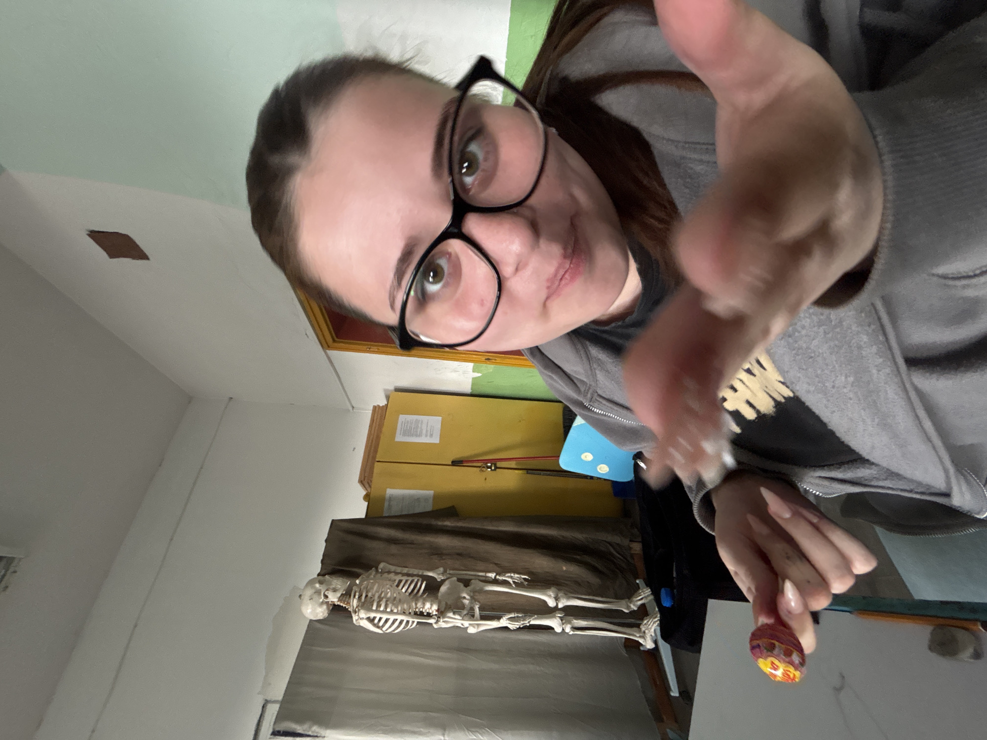 The image depicts an indoor scene, likely a classroom or laboratory, located in the city of Troyan, Bulgaria. In the foreground, a young female with brown hair and black-rimmed glasses faces the camera, looking directly at it with a slight smile. She wears a grey hooded sweatshirt over a dark t-shirt and has long, light-colored fingernails. Her right hand is extended, partially blurred and open towards the camera, while her left hand holds a red and yellow lollipop.
In the mid-ground to the left, a full-sized human skeleton model stands upright on a base. Behind the skeleton, a large, dark brown fabric curtain or drape covers part of the wall. To the right of the skeleton and curtain, a tall, yellow two-door storage cabinet is visible, with several sheets of paper taped to its right door. Leaning against the cabinet are various objects, including a red stick-like item and a black stick-like item. A light blue object, possibly a small chair or board, is positioned near the cabinet's base. Further right, a white wall with a light green lower section forms a corner. A brown wooden door frame with yellow trim is partially visible behind the female. The ceiling is white, with a visible vent or fixture. A white surface, possibly a desk or drawing board, along with a yellow ruler and other small objects, are visible in the lower left foreground. The walls show minor imperfections.