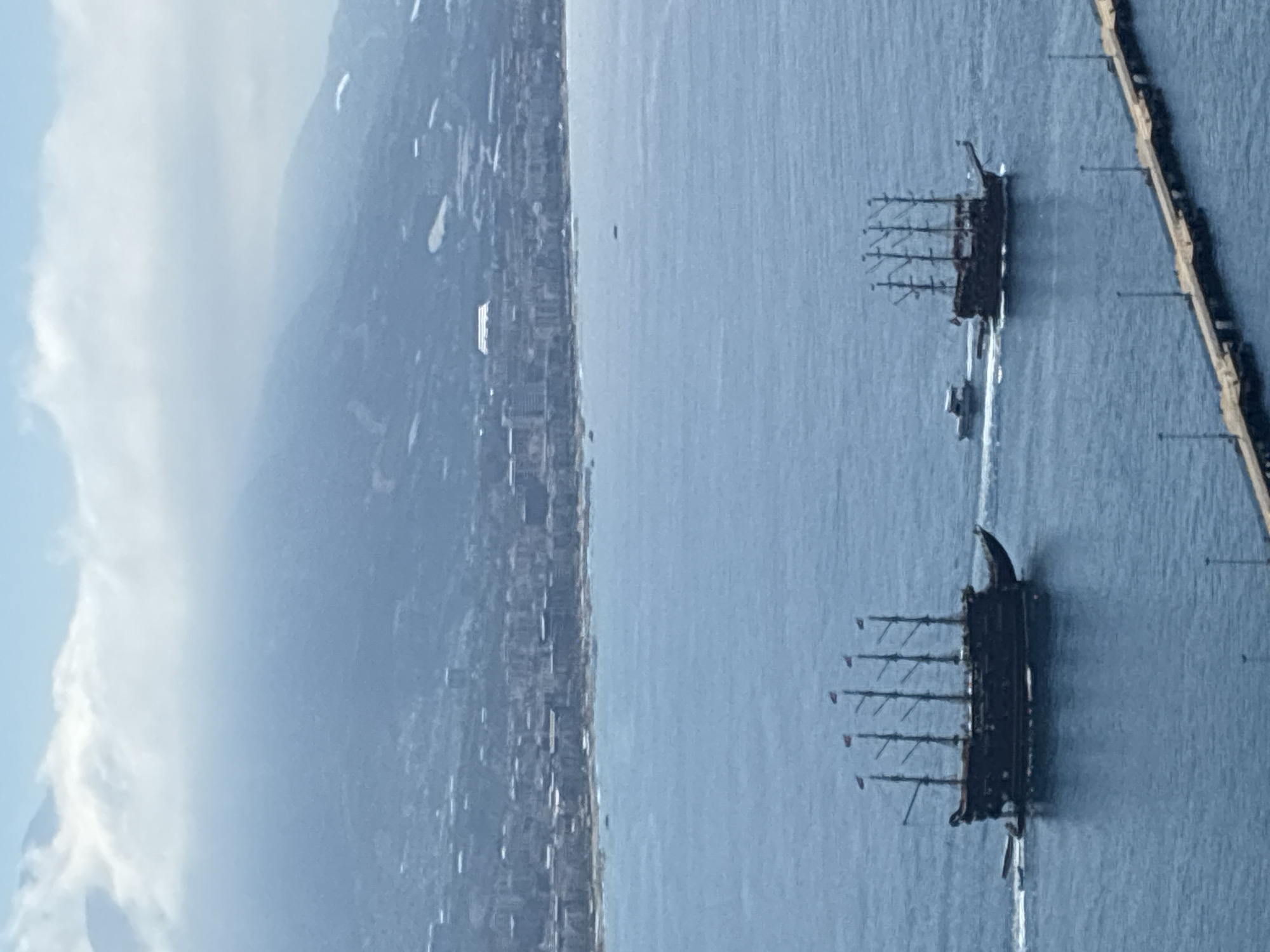 The image captures a wide aerial view of a bustling coastal scene, likely Alanya, Turkey, featuring maritime activity against a mountainous backdrop.
In the foreground and midground, two large, dark-hulled ships, resembling multi-masted pirate ships or historical sailing vessels, traverse the calm blue water. Each ship leaves a distinct white wake behind it, indicating motion. One ship is closer to the bottom right, and the other further up and to the right. A smaller, modern powerboat is visible between the two larger vessels, also in motion. Along the far right edge of the frame, a long, light-colored pier or breakwater extends into the water, dotted with what appear to be streetlights or pylons.
The midground reveals a densely populated city or town stretching along the coastline, with numerous buildings clustered at the base of prominent mountains that dominate the left and central background. The city's structures appear to be light-colored against the darker hues of the landscape. The mountains are large and imposing, fading into a hazy, blue-gray mist towards their peaks.
The sky occupies the upper portion of the image, displaying a mix of clear blue and soft, white clouds, particularly noticeable on the left side, suggesting a partly cloudy day. The overall lighting indicates daytime, likely late morning or early afternoon, with bright but diffused light. No specific text is discernible anywhere in the image.