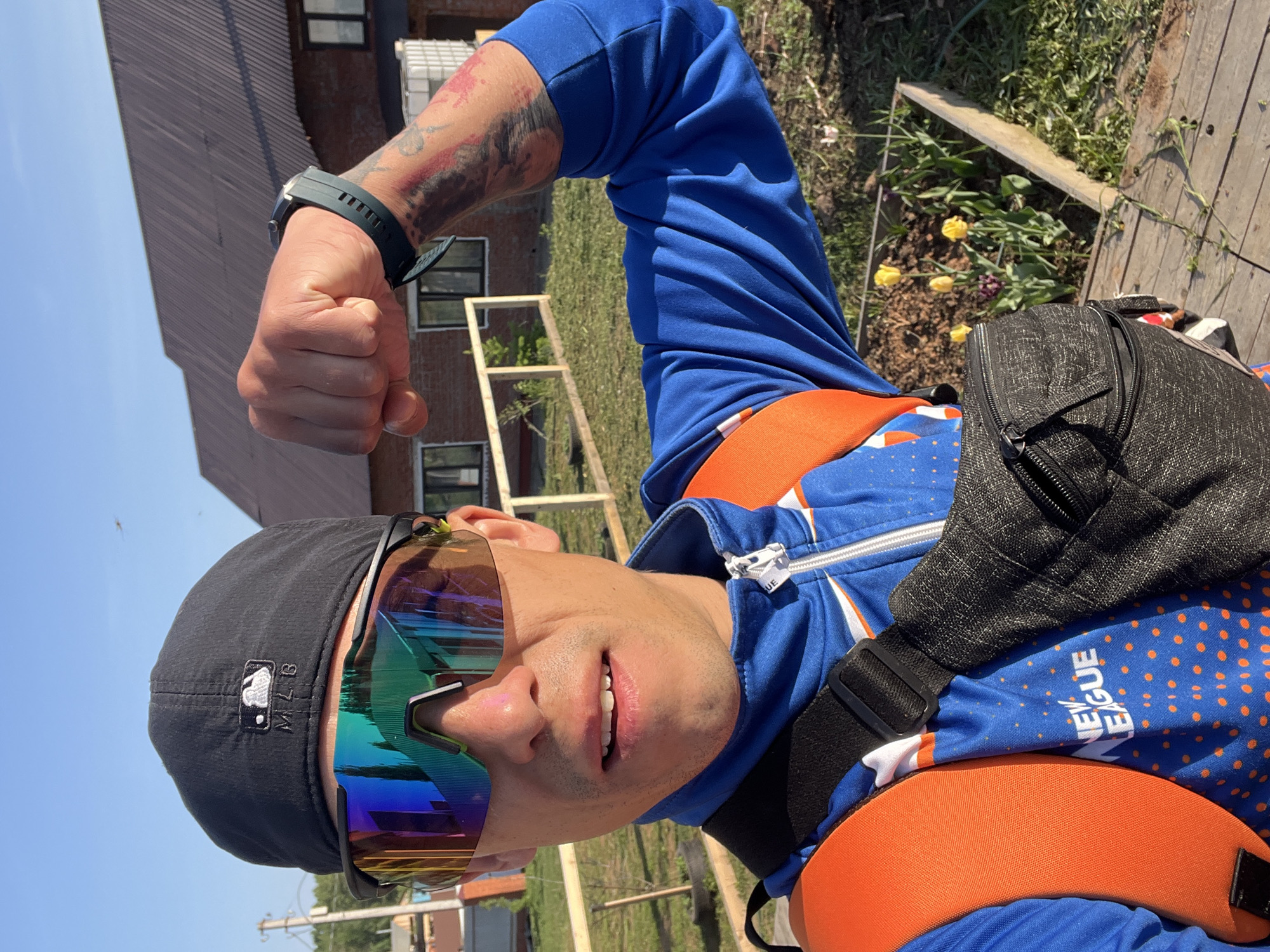 A man is posing outdoors with his fist raised. He is wearing a black baseball cap with an MLB logo, mirrored sunglasses, a blue zip-up jacket with orange accents, and a black fanny pack. The man has a tattoo on his left arm and a dark watch on his left wrist. Behind him is a building, some wooden structures, and a patch of grass with flowers. The location is likely Chaikovsky, Russia.