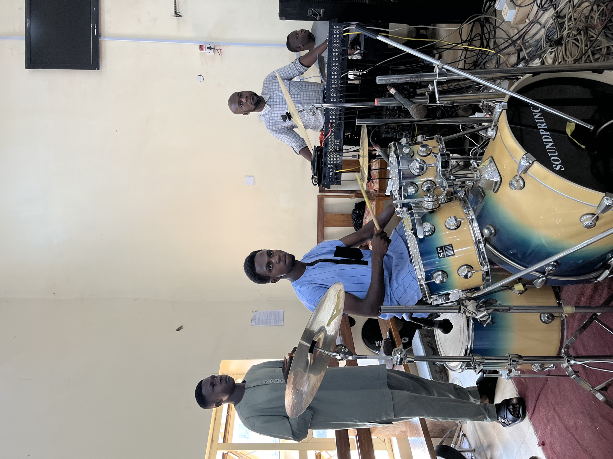 The image shows three men playing musical instruments indoors. A man in a light green long-sleeved shirt holds a cymbal. A man in a blue-striped shirt plays a drum set. A third man, wearing a checkered shirt, stands behind the drum set and appears to be playing a keyboard or mixing console. Various cables and wires are visible near the keyboard. The drum has "SOUNDPRINC" visible on the side. The location is possibly a church, based on the style of dress, however, there is insufficient information in the image to determine the location.
