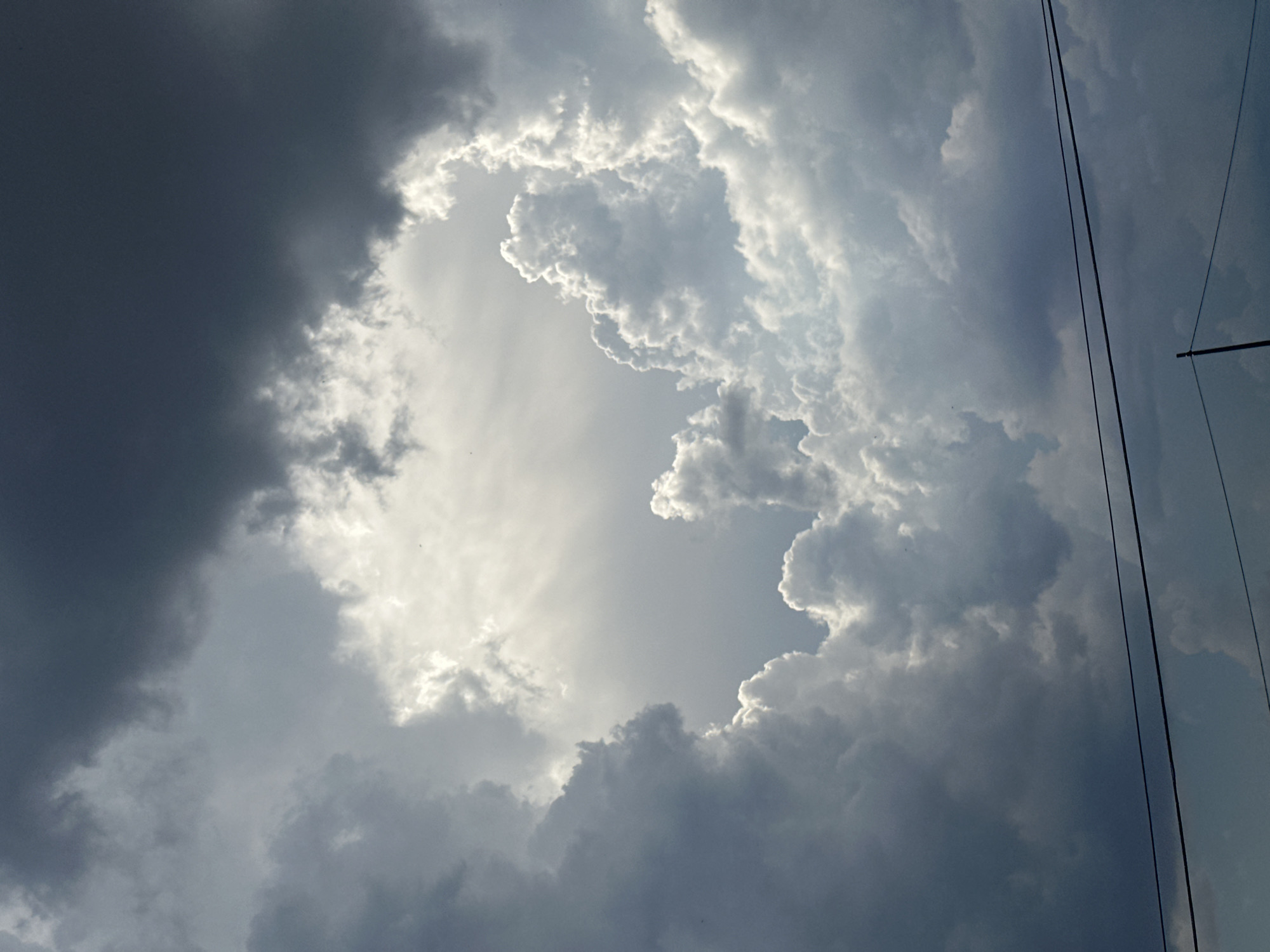 The image depicts a dramatic sky filled with dark, heavy storm clouds and bright, illuminated clouds, creating a contrast that forms a rough, oval-shaped opening. The bright clouds are puffy and textured, catching the sunlight which streams through the opening, illuminating the scene from above. The dark clouds surround this bright area, appearing dense and foreboding. In the foreground, two thin, dark wires run horizontally across the lower portion of the image, suggesting the presence of a power line or telephone line. Below the wires, a thin, vertical pole is visible on the right side of the frame. The overall impression is of an impending storm with a break in the clouds allowing sunlight to pierce through. The image does not contain any elements that would specifically place it in the city of Assisi, Italy; it is a generic sky scene.