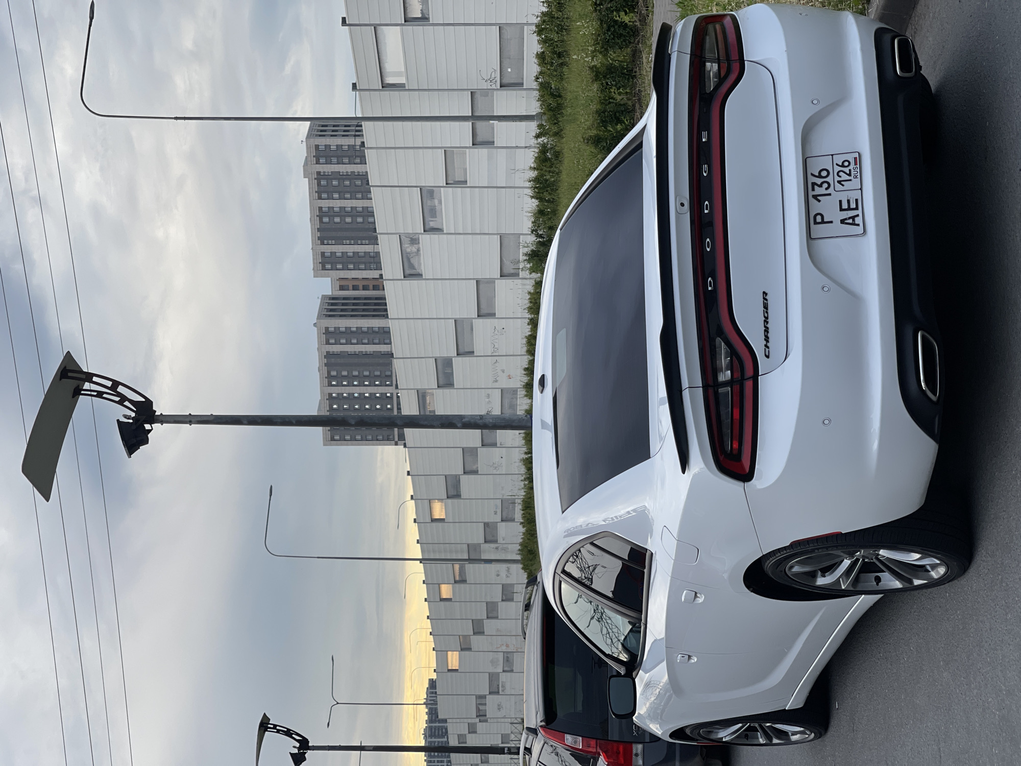 A white Dodge Charger is parked on a paved road in an urban setting. The car is viewed from the rear and right side, showcasing its taillights, a spoiler, and the rear bumper with dual exhaust pipes. A license plate with Russian markings, "P 136 AE 126 RUS", is visible. The car is parked next to a dark-colored vehicle, possibly an SUV. In the background, a tall, white sound barrier wall runs along the road, with square panels and some graffiti. Behind the wall, several tall residential buildings with gray facades and numerous windows are visible. Streetlights with a modern design stand along the road. The sky is overcast, with some diffused light suggesting either early morning or late afternoon. Green vegetation is present along the base of the sound barrier. The environment appears to be a city or suburban area with active construction or recent development, indicated by the modern buildings and infrastructure.