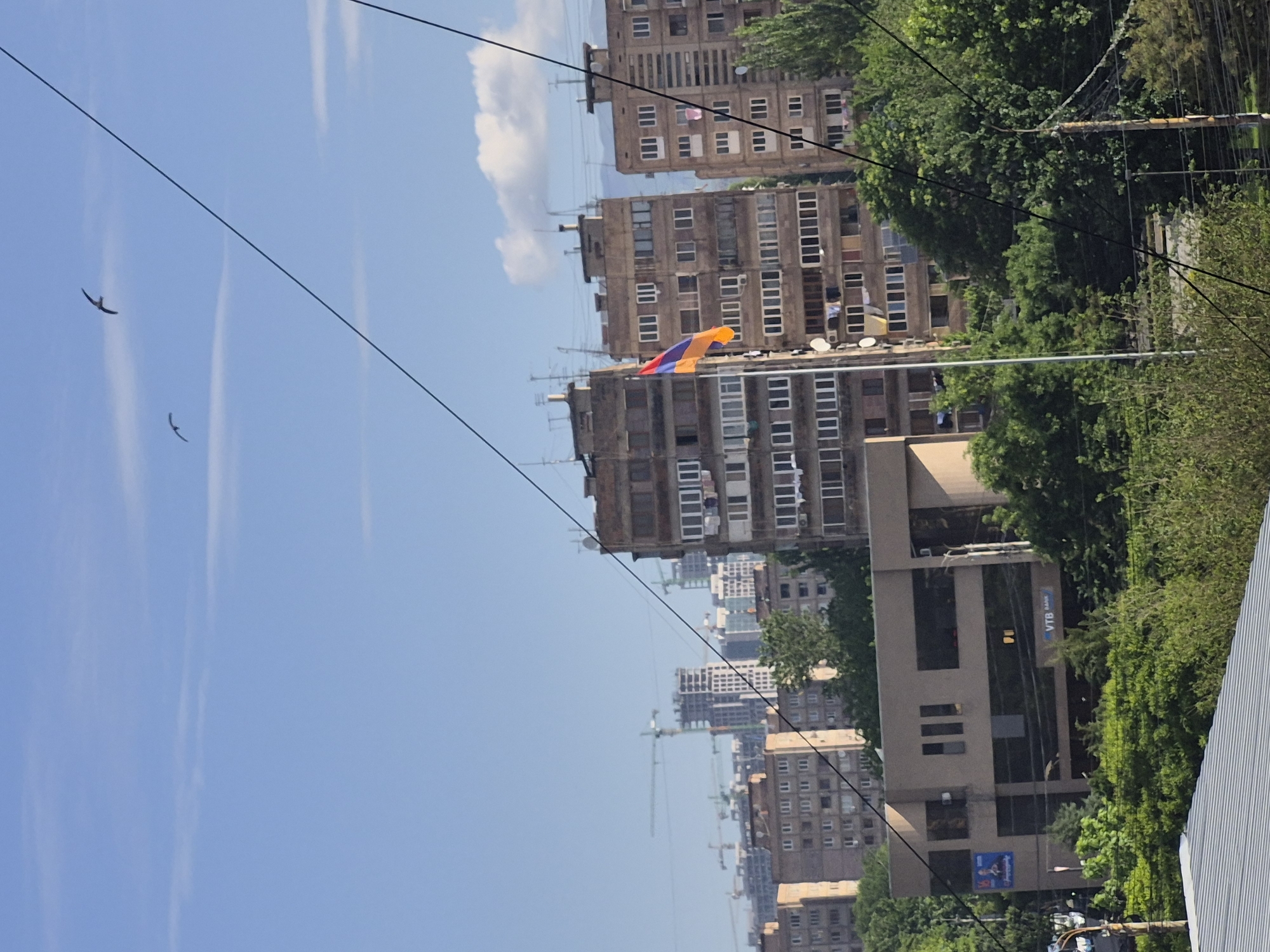 This media file presents an urban landscape in Yerevan, Armenia, captured on a clear, bright day. The sky, a vivid blue with a few faint cirrus clouds, occupies the upper left section of the frame. Two small, dark birds are visible in flight against the sky. Several thin, dark wires, likely power lines or communication cables, traverse the image diagonally.
The mid-ground and background are dominated by multi-story buildings, typical of Soviet-era or post-Soviet architecture, featuring numerous windows and some balconies. On the roof of one building, the national flag of Armenia, with its distinctive red, blue, and orange horizontal stripes, is proudly displayed.
Green trees and dense foliage are abundant on the right side of the image, interspersing with and partially obscuring the urban structures. In the far distance, the presence of several construction cranes indicates ongoing development within the city.
In the lower right, a modern building with prominent signage is visible. The text "VTB Bank" is clearly legible on its facade, and a smaller "VTB" logo appears nearby. A blue banner or billboard below also shows the text "Viva-MTS". The scene depicts a lively city environment under a sunny sky.