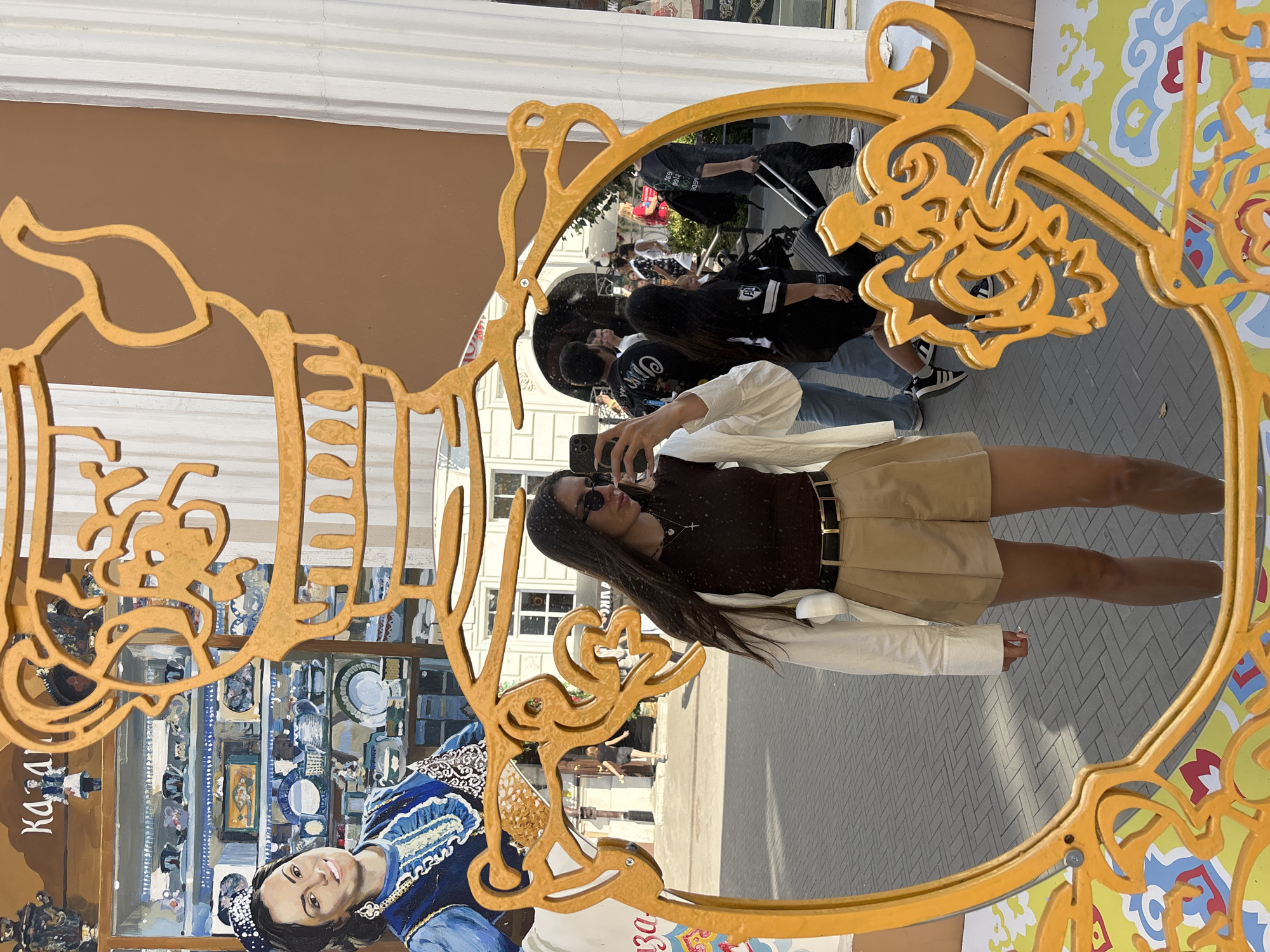 The image captures a street scene reflected in a decorative, golden, ornate mirror frame. In the foreground, a young woman is taking a selfie, holding a smartphone in her right hand. She is wearing a brown tank top, a white unbuttoned shirt, khaki shorts, a black belt, and sunglasses. She has long dark hair and is wearing a delicate necklace with a cross pendant.
The mirror reflects a bustling street with several people walking. The background of the reflection shows buildings with white columns and windows, suggesting an urban environment. The backdrop behind the woman, not in the mirror's reflection, features a mural depicting traditional Russian imagery. Specifically, the mural includes a stylized depiction of a woman in traditional attire, shelves filled with porcelain dishes and samovars, and the word "КАZАНЬ" (KAZAN) in Cyrillic letters. This mural indicates the location is in Kazan, Russia.
The overall scene suggests a tourist or leisure activity, with the woman posing for a photo in front of a culturally themed backdrop. The time of day appears to be daytime, with bright sunlight casting shadows on the ground.