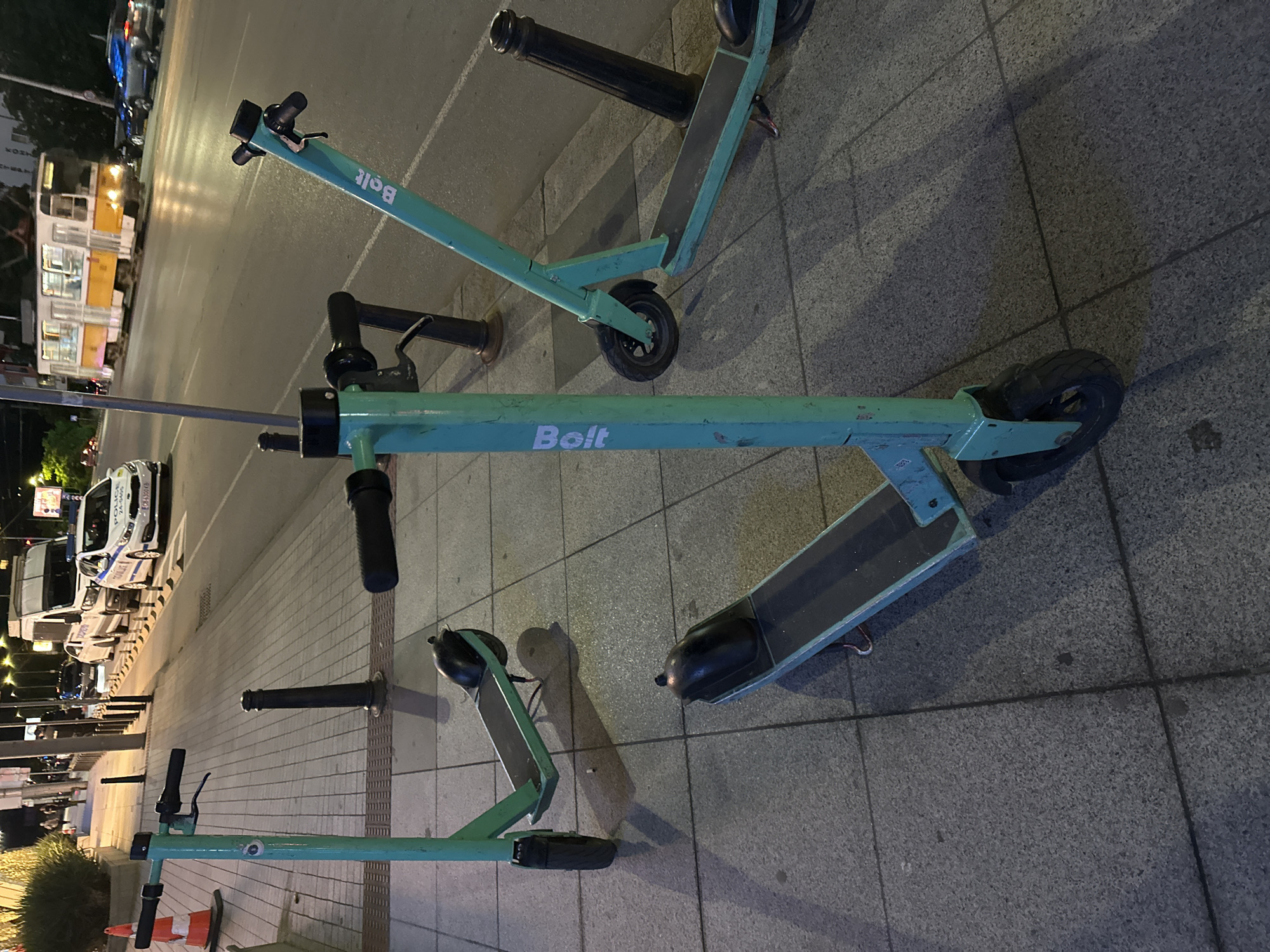 In Sofia, Bulgaria, on a sidewalk with gray stone tiles, three teal-colored electric scooters are parked. One scooter is in the foreground, facing left, with its handlebar and front wheel visible. Behind it and to the right, another scooter is partially visible, angled away from the camera. To the left and further back, a third scooter is parked, also angled away. The scooters have the "Bolt" logo on their frames. The sidewalk is bordered by a street with traffic. In the background, a white van, a police car, and a yellow tram are visible. Streetlights illuminate the scene.