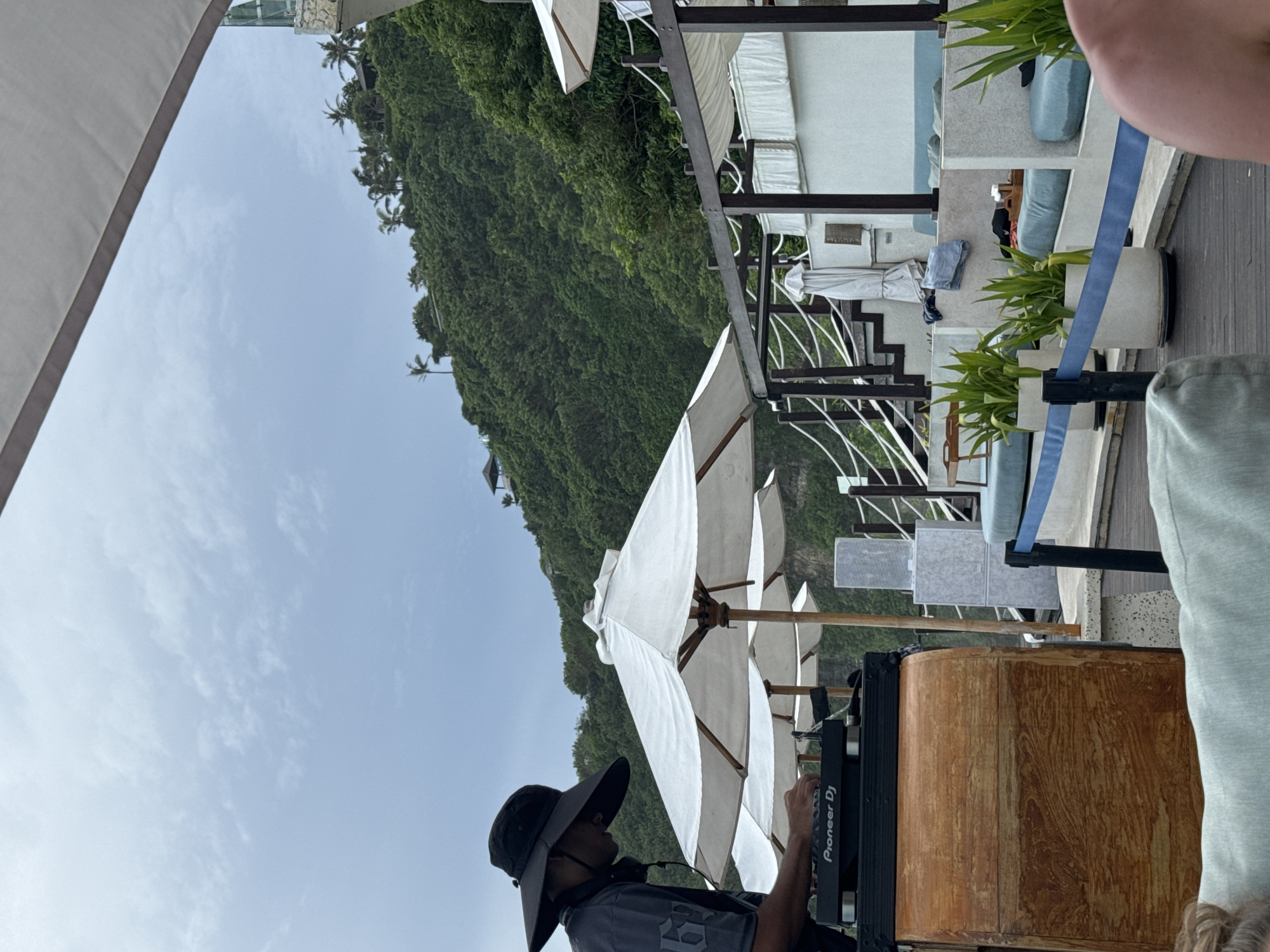 This image depicts an outdoor, luxurious resort or beach club setting in Kuta Selatan, Indonesia, captured during bright daylight hours under a clear to partly cloudy sky.
The scene is anchored by a steep, lush green hillside or cliff covered in dense vegetation, rising prominently in the background. Scattered palm trees and some white modern-style buildings or structures can be seen on the upper slopes of the hill.
In the mid-ground and foreground, a multi-level patio or deck area features white, cushioned sun loungers and comfortable seating. Several large white parasols are open, providing shade. The flooring appears to be dark wooden decking. Potted green plants add to the natural aesthetic, and a distinctive blue strap or ribbon functions as a decorative barrier along the edge of the patio.
A central figure is a person, likely a DJ, positioned on the right side of the image. They wear a wide-brimmed black hat and a dark shirt, with headphones visible around their neck. They are actively operating professional DJ equipment, which clearly displays the text "Pioneer DJ" on its front panel. The equipment rests on a rustic wooden cabinet or stand. Part of another person's body, possibly an arm or leg clad in light-colored fabric, is visible on the far right, suggesting other patrons are present.
The overall impression is one of relaxation and leisure in an upscale tropical environment, possibly with music being played.