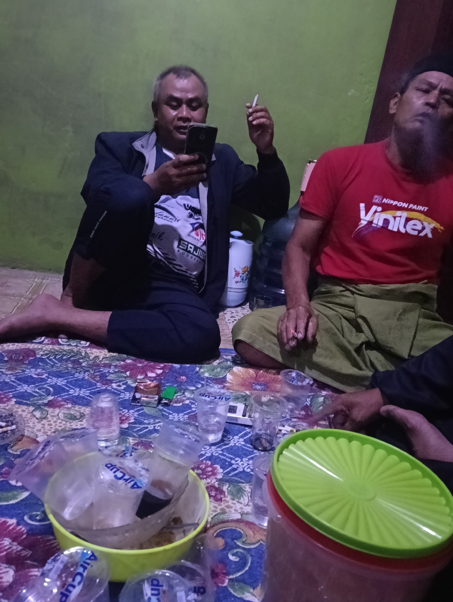 The image depicts two adult males seated on a patterned rug indoors, likely in Tasikmalaya, Indonesia, given the cultural context implied by the clothing and setting. The room appears dimly lit, with a green wall in the background.
The man on the left is wearing a dark jacket over a white and black striped shirt. He is holding a black smartphone in his right hand and is looking at the screen. A lit cigarette is held between the index and middle fingers of his left hand. His legs are crossed, and his bare feet are visible.
The man on the right is wearing a red t-shirt with a "Nippon Paint Vinilex" logo and a green sarong. He has a beard and is exhaling smoke from a cigarette he holds between his fingers. His legs are also crossed, and he is seated next to a large water dispenser.
Scattered on the rug are several plastic cups, some containing liquid, and cigarette packs, indicating a casual gathering or social interaction. A large green-lidded container is also visible in the foreground. The overall atmosphere suggests an informal social setting.