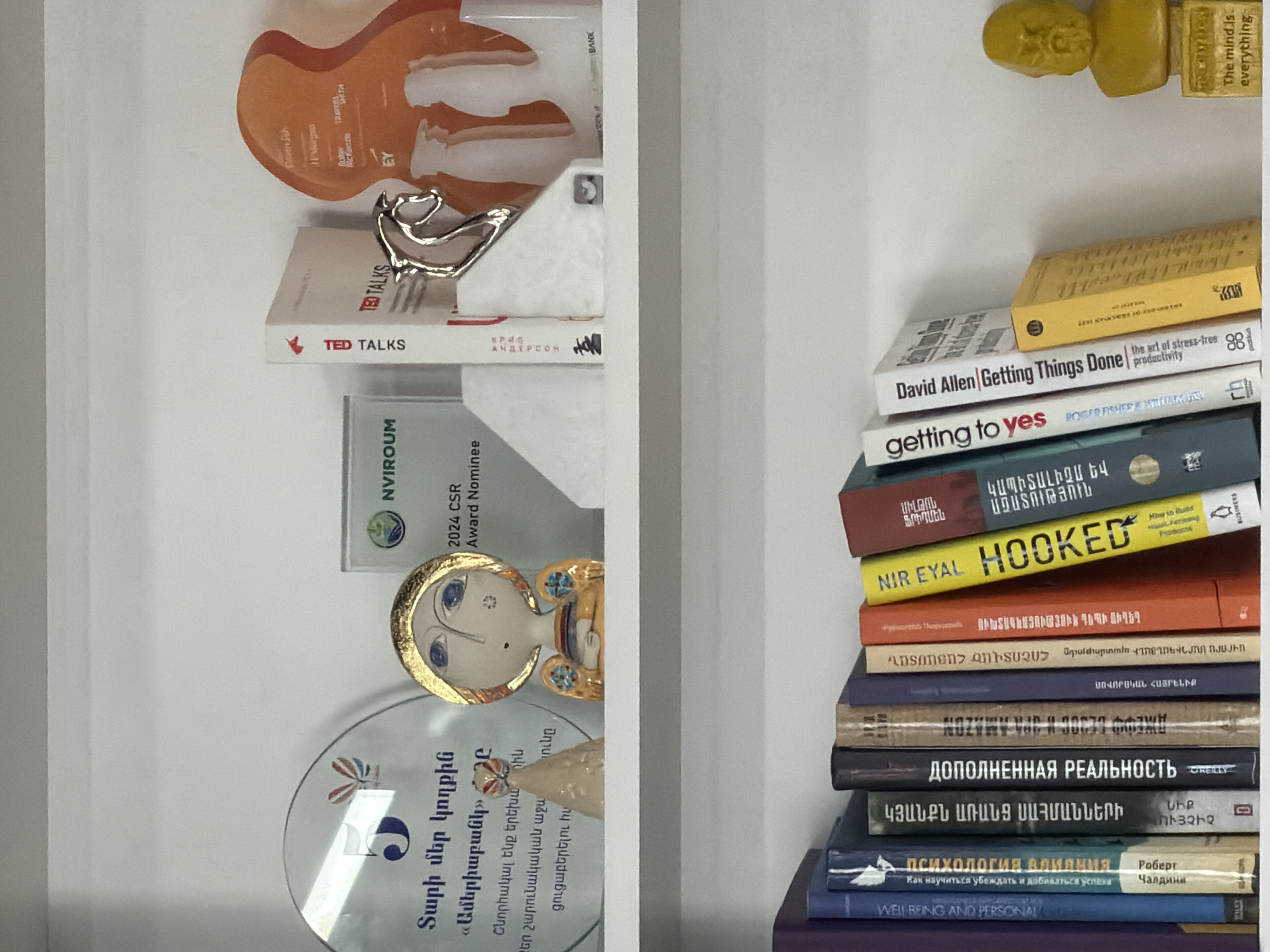The image shows a bookshelf with various items and books, likely located in Yerevan, Armenia, based on the presence of Armenian text on the books and awards.
On the top shelf, there are several awards: an orange award trophy, a silver bird sculpture, a "TED Talks" book, and a "NVIROUM 2024 CSR Award Nominee" plaque. There's also a small decorative item with a face.
The bottom shelf contains a stack of books in Armenian and English, along with a small golden sculpture. The English books include "David Allen - Getting Things Done", "Getting to Yes", and "Hooked" by Nir Eyal.