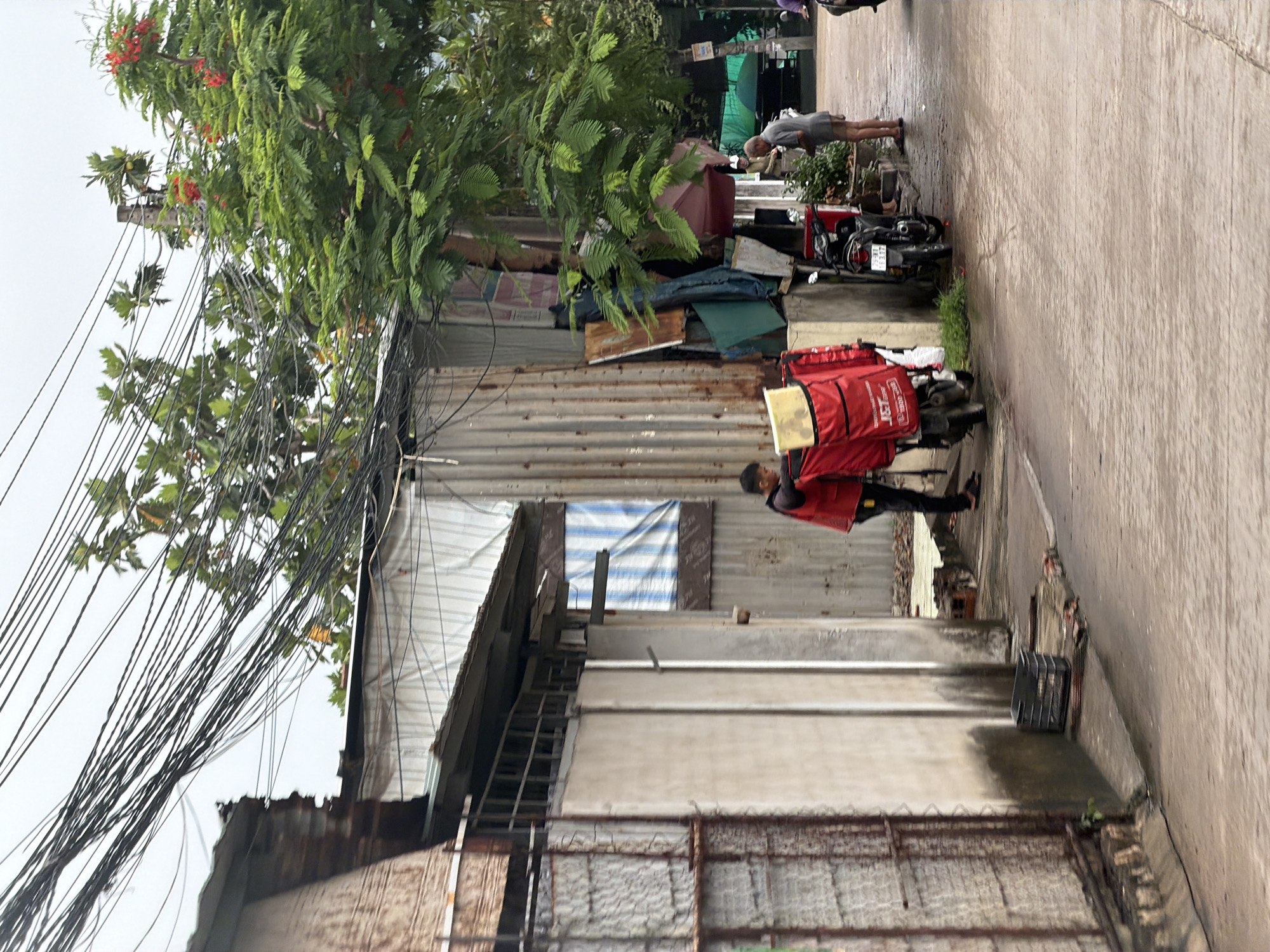 The image depicts a street scene in Nha Trang, Vietnam, characterized by a narrow roadway and a row of buildings with corrugated metal and plastered walls. Overhead, a dense network of electrical wires crisscrosses the sky, common in urban Vietnamese settings. In the foreground, a person wearing a red vest with the "JET" logo and black pants stands next to a motorcycle equipped with a large red delivery box. Another motorcycle is parked nearby. To the right, an older man in a grey shirt and shorts stands on the sidewalk. The overall environment suggests a mixed-use area with residential or small commercial structures and evidence of ongoing daily activities, such as deliveries. The ground is wet, indicating recent rain.