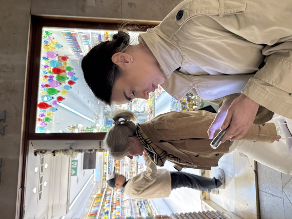 The image displays a street scene in Venice, Italy, positioned outside a Murano glass shop. Three individuals are visible. In the right foreground, a young adult female with dark hair styled in two low buns, wearing a light beige trench coat, holds a smartphone with both hands, its screen illuminated, and looks down at it. A white shopping bag is partially visible held in her left hand. Behind her, an older adult female with blonde hair in two ponytails, wearing a tan jacket and a plaid scarf, faces left and looks into the shop's interior. In the background, inside the brightly lit shop, an adult male in a beige jacket, dark pants, and a white baseball cap stands, facing away from the viewer, observing items on a display shelf. The shop's windows and open entrance reveal extensive displays of colorful Murano glass objects, including figurines, jewelry, and decorative items. The upper section of the right storefront window features an arrangement of multiple small, multicolored glass hot air balloon ornaments. The building facade above the shop entrance consists of textured, light-colored stone or stucco.