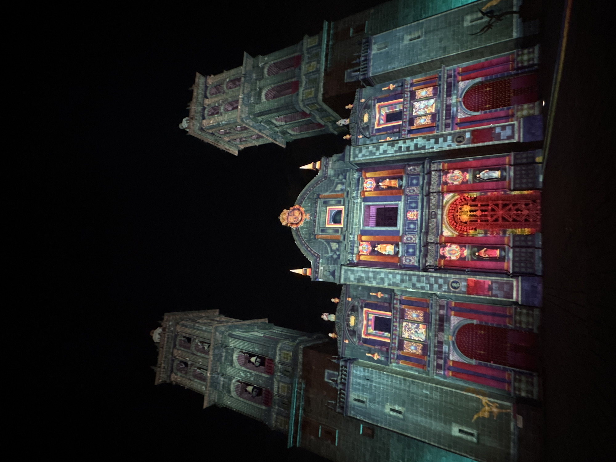 The image shows the Cathedral of Puebla in Puebla, Mexico, at night. The cathedral is illuminated with colorful lights and projections, highlighting its architectural details. The building has two towers on either side of the main facade, which is elaborately decorated with statues and carvings. The night sky is dark.
