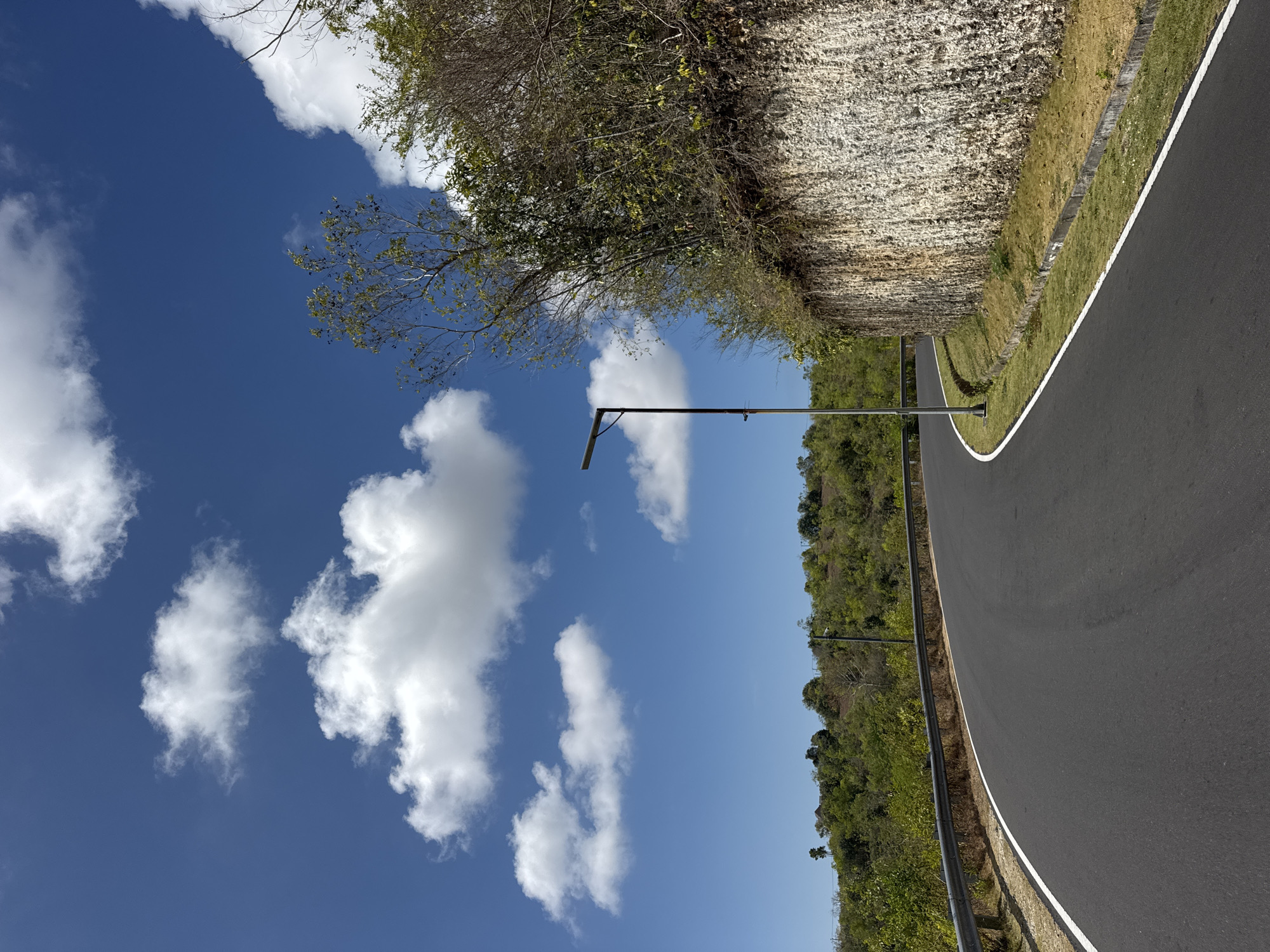 The image depicts a paved, curved road in Kuta Selatan, Indonesia. The road features white lane markings and is bordered by a metal guardrail on the left and a steep, rocky embankment on the right. A single modern streetlamp stands near the road. The landscape consists of lush green hills covered in dense vegetation, including trees and shrubs. The sky is a vibrant blue, dotted with scattered white cumulus clouds. The scene appears to be captured during daylight, with clear weather conditions.