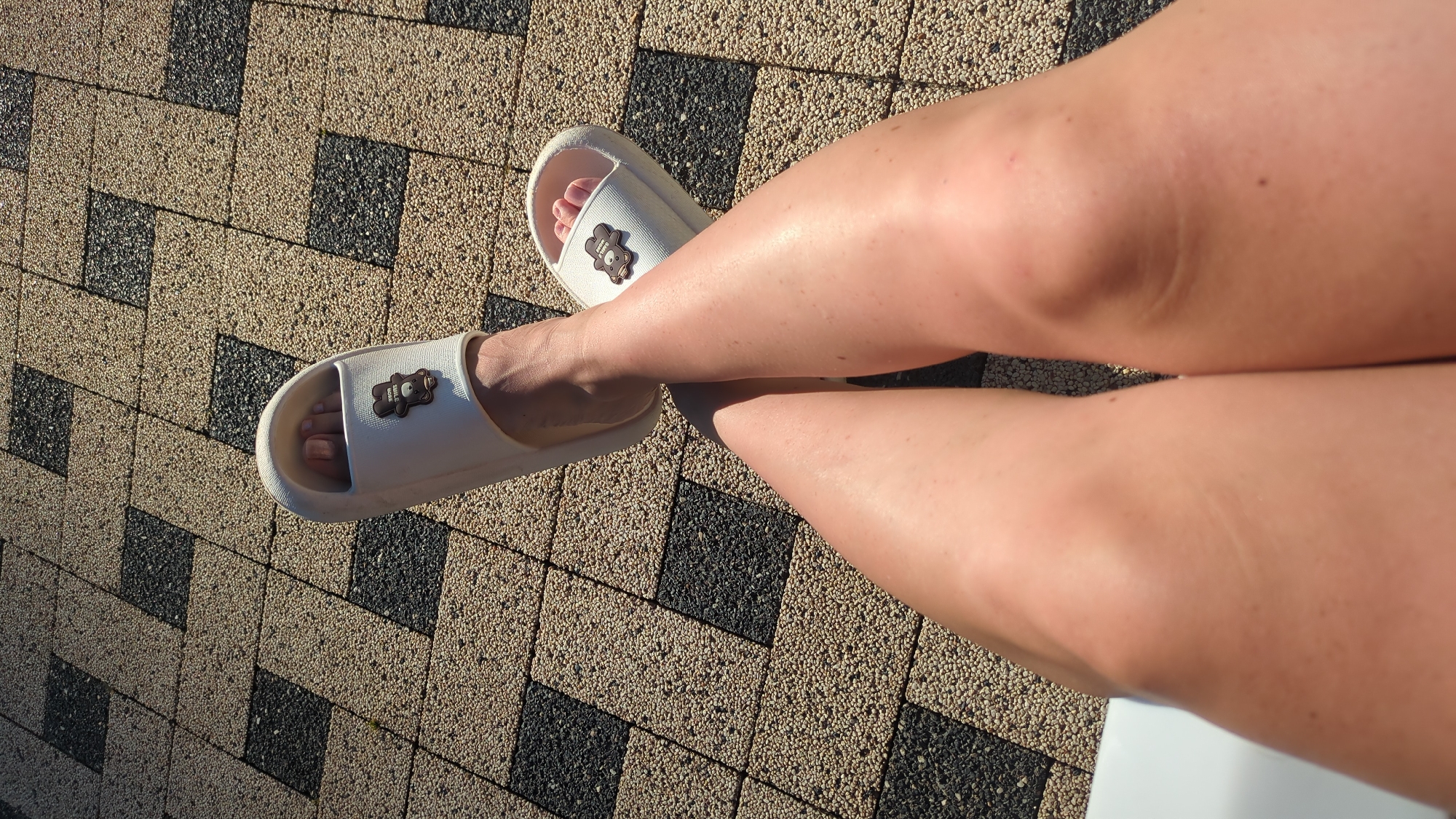 The image shows a pair of white slide sandals with a small teddy bear charm on each. The sandals are worn by a person whose legs and feet are visible. The person's legs are crossed, and they are resting on a tiled surface. The tiles are arranged in a geometric pattern of alternating light brown and dark grey squares. The lighting suggests it is daytime, possibly outdoors. The specific location is a tiled patio area in Vadul lui Voda, Moldova.