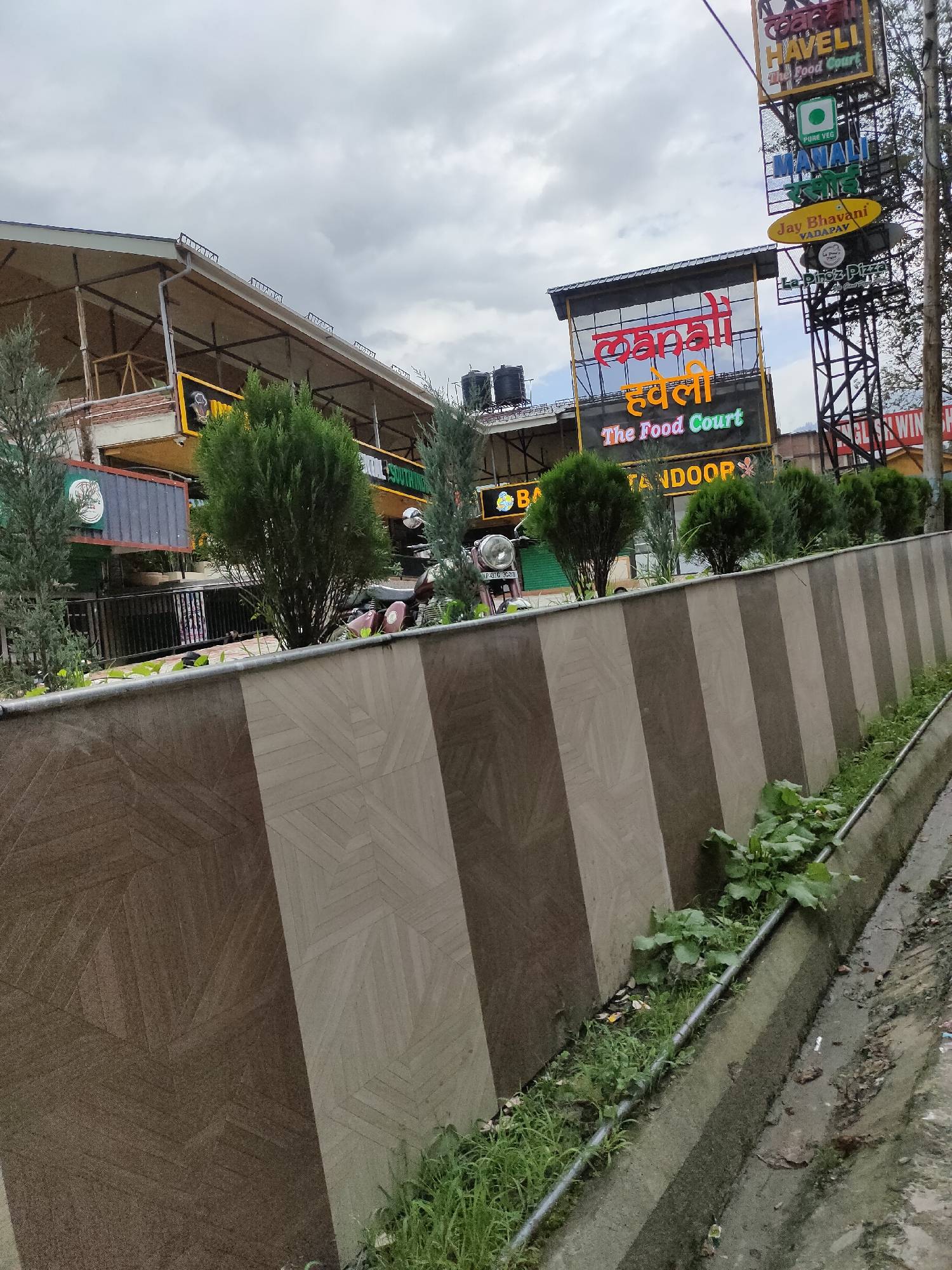 The image shows a street-level view of "Manali Haveli The Food Court" in Bahang, India. A large, multi-tiered sign for the establishment is visible on the right side of the image. The sign displays "Manali Haveli" in large Hindi script, followed by "The Food Court" in English. Below this, smaller signs indicate "Tandoor" and "Jay Bhavani Vada Pav". The establishment appears to be a food court with various dining options. In the foreground, a concrete barrier with a patterned design separates the street from the food court. Vegetation, including grass and weeds, grows along the edge of the barrier and in a drainage ditch. The sky is overcast. A motorcycle is parked near the entrance of the food court. The overall impression is of a commercial area in a town or city.