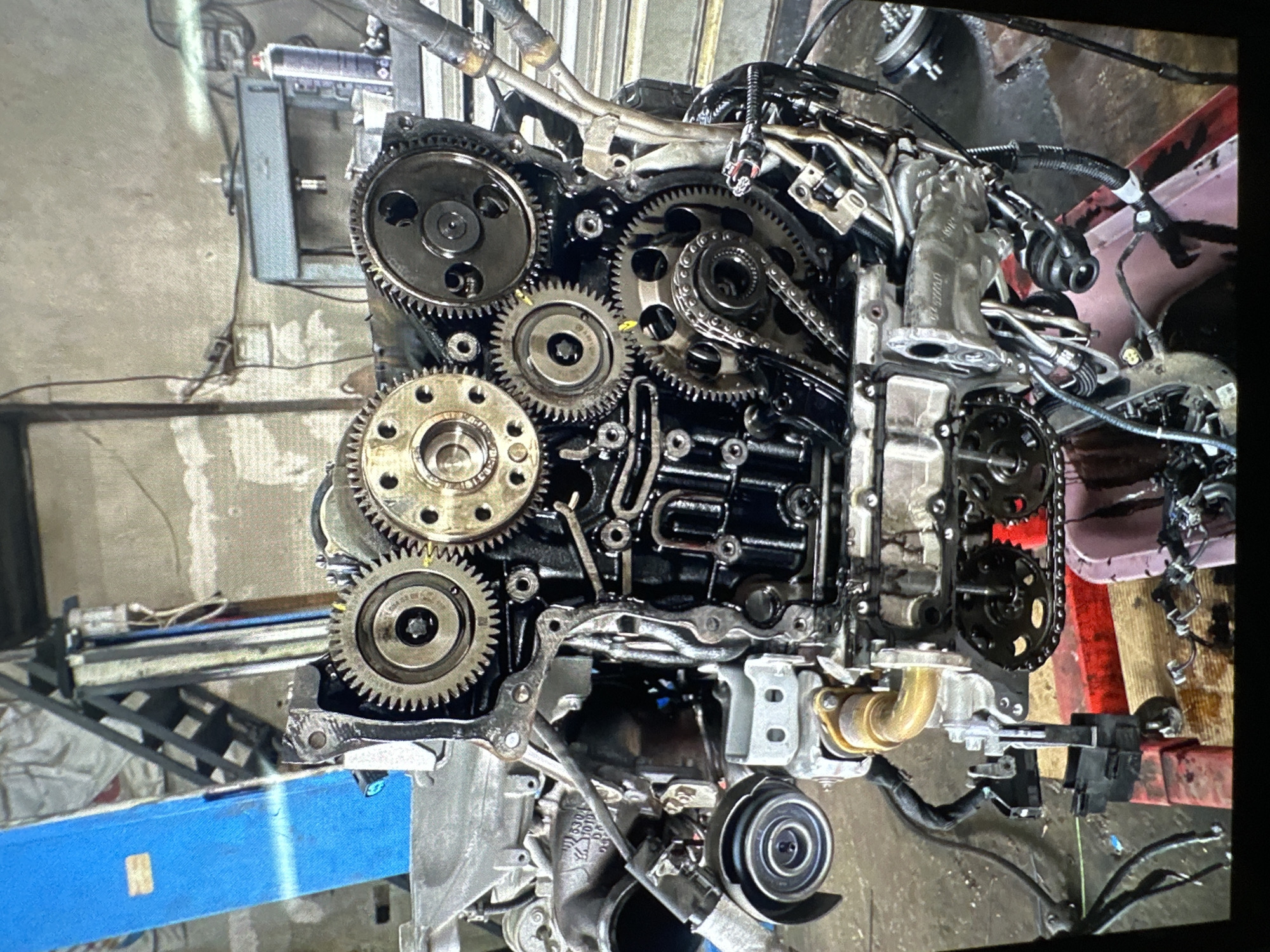 The image displays a close-up view of an internal combustion engine, specifically the timing gear and chain assembly, undergoing maintenance or repair in what appears to be an automotive workshop. The engine block is positioned vertically, with its timing cover removed, exposing a complex arrangement of metallic gears and a timing chain.
The central focus is on several large and small gears, some with visible teeth, and a dark metal timing chain that connects them. The gears are various shades of metallic grey and bronze, with some exhibiting oil residue and signs of wear. The engine block itself is dark, likely from engine oil. Various bolts, fasteners, and internal passages of the engine are visible within the block.
In the background, to the left, parts of workshop equipment are visible, including a blue hydraulic lift or stand. To the upper left, a grey metal workbench or another piece of machinery can be seen. The floor beneath the engine appears to be concrete. To the right of the engine, several wires, hoses, and tubes, part of the engine's ancillary systems, are tangled together. Towards the lower right, a component resembling a turbocharger or part of the exhaust system is attached to the engine block. At the bottom center, a pulley or tensioner for a serpentine belt is visible.
Notable details include oil sheen on many of the engine's internal components, indicating a working engine that has been disassembled. On one of the larger gears, faint text is visible, possibly including "MADE IN GERMANY" and a part number, although it is mostly illegible due to resolution and angle. Another gear also shows some embossed numbers like "030 109 088 B". The scene captures an active work environment in Chirpan, Bulgaria, focused on automotive mechanics.