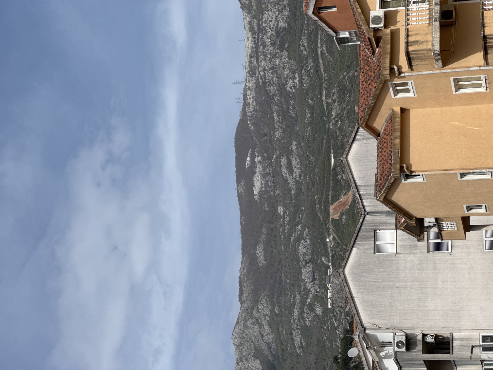 The image shows a view in Budva, Montenegro, looking towards a rocky mountain. Buildings with tile roofs and light-colored facades are in the foreground on the right. The sky is partly cloudy. Overhead power lines are visible on the mountain slope.