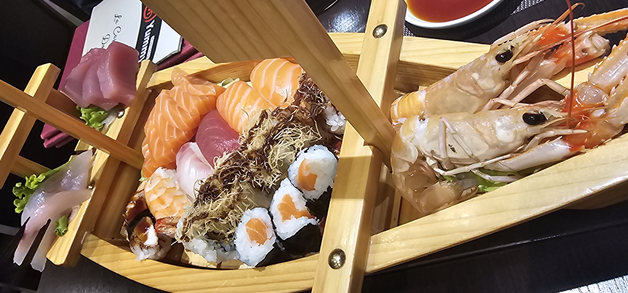 A wooden sushi boat holds an assortment of raw fish and seafood. The top tier displays a few pieces of white fish and a pile of pink tuna sashimi. Below, the boat is filled with various sushi rolls, sashimi slices of salmon and tuna, and what appears to be shredded seaweed or a similar garnish. Two large, raw langoustines with prominent antennae and segmented bodies rest on a bed of lettuce in the lower section of the boat. A small dish of red dipping sauce is visible to the right of the boat. The setting appears to be a restaurant.