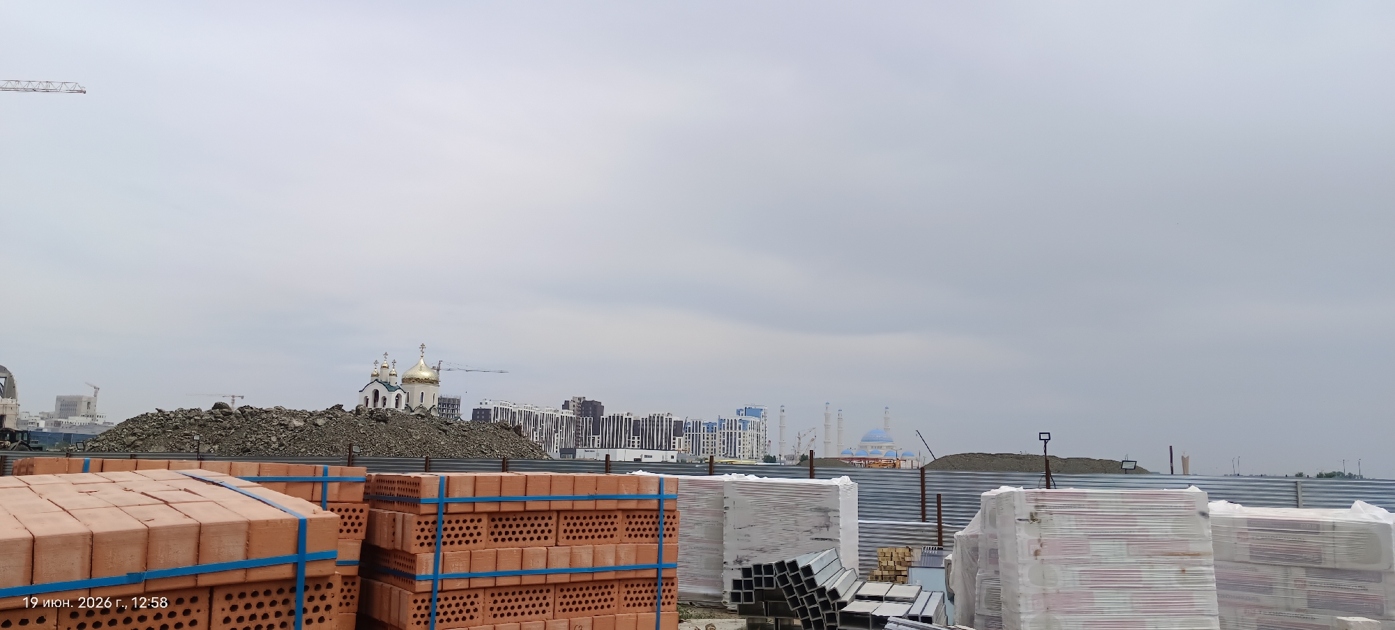 The image depicts a construction site in Astana, Kazakhstan, with stacks of bricks in the foreground. In the background, several buildings are visible, including a church with golden domes, a mosque with blue domes, and a cluster of modern apartment buildings. The sky is overcast, and construction cranes can be seen in the distance, indicating ongoing development. The timestamp in the lower left corner reads "19 июня 2026 г., 12:58," which translates to "June 19, 2026, 12:58." The scene is a typical urban construction environment under a cloudy sky.