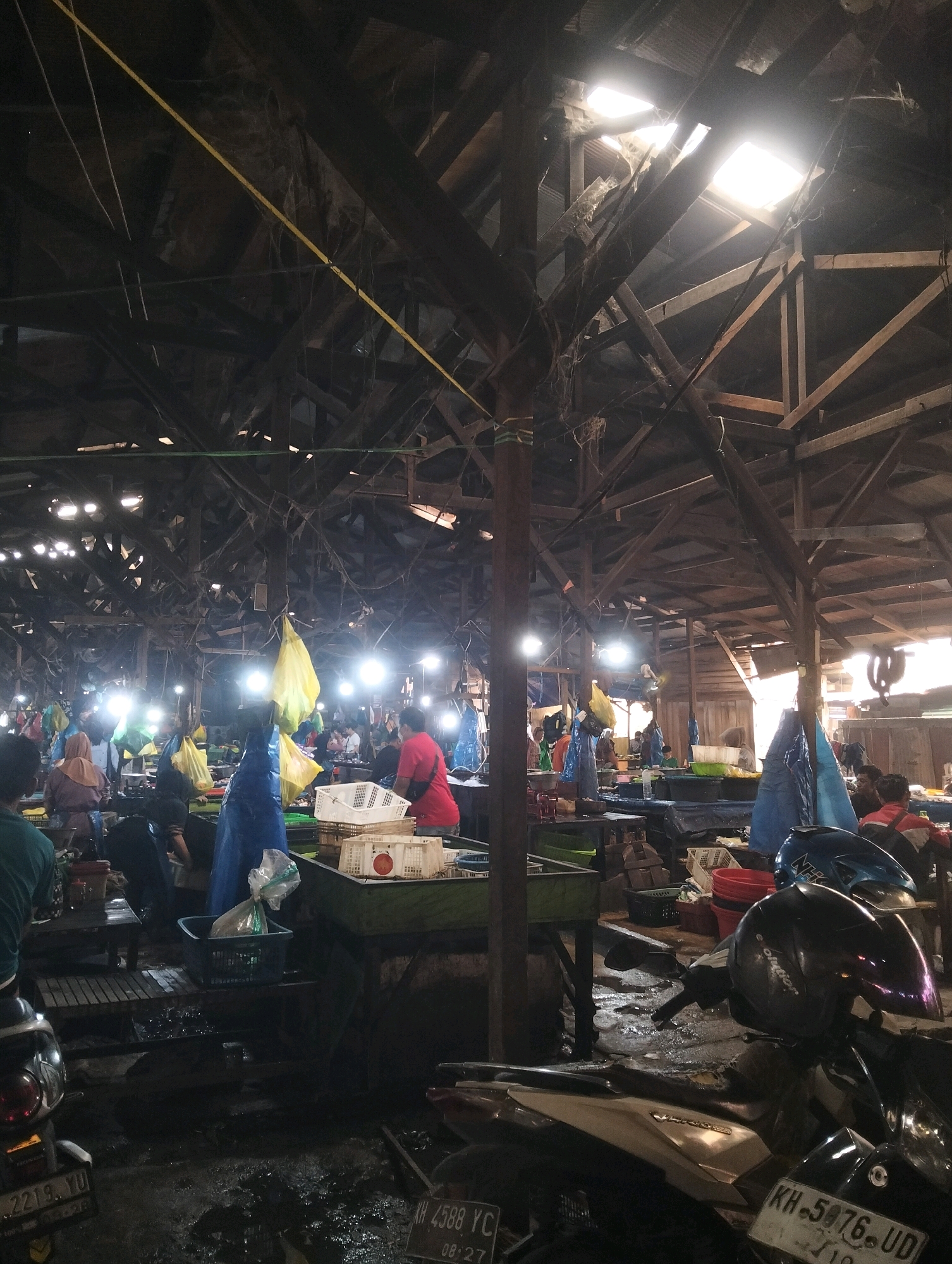 This image depicts the bustling interior of a traditional market in Palangka Raya, Indonesia, likely a fish market given the presence of numerous blue plastic sheeting covers, white plastic crates, and what appear to be buckets, all common for handling seafood. The market's structure is rustic, with a weathered wooden beam ceiling and exposed wooden supports, some of which are laden with cobwebs. Natural light filters through gaps in the roof, creating bright patches, while artificial lights are also visible, illuminating the scene and casting shadows.
In the foreground, several motorcycles are parked, their surfaces wet and reflective, indicating a damp or recently rained-on environment. One motorcycle in the center is equipped with two helmets, one black and one blue, suggesting its owner is present or has just left.
The middle ground of the market is a hive of activity. People, some wearing headscarves or hats, are engaged in various tasks, seemingly tending to stalls or interacting with customers. Colorful plastic bags, some yellow and some green, hang suspended from the ceiling or are placed on stalls, possibly containing goods for sale. A person in a red shirt is prominently visible, appearing to be a vendor.
The overall atmosphere is one of commerce and daily life in a crowded marketplace. The floor is wet and somewhat uneven, typical of such environments. The composition captures the raw, unpolished reality of a local Indonesian market.