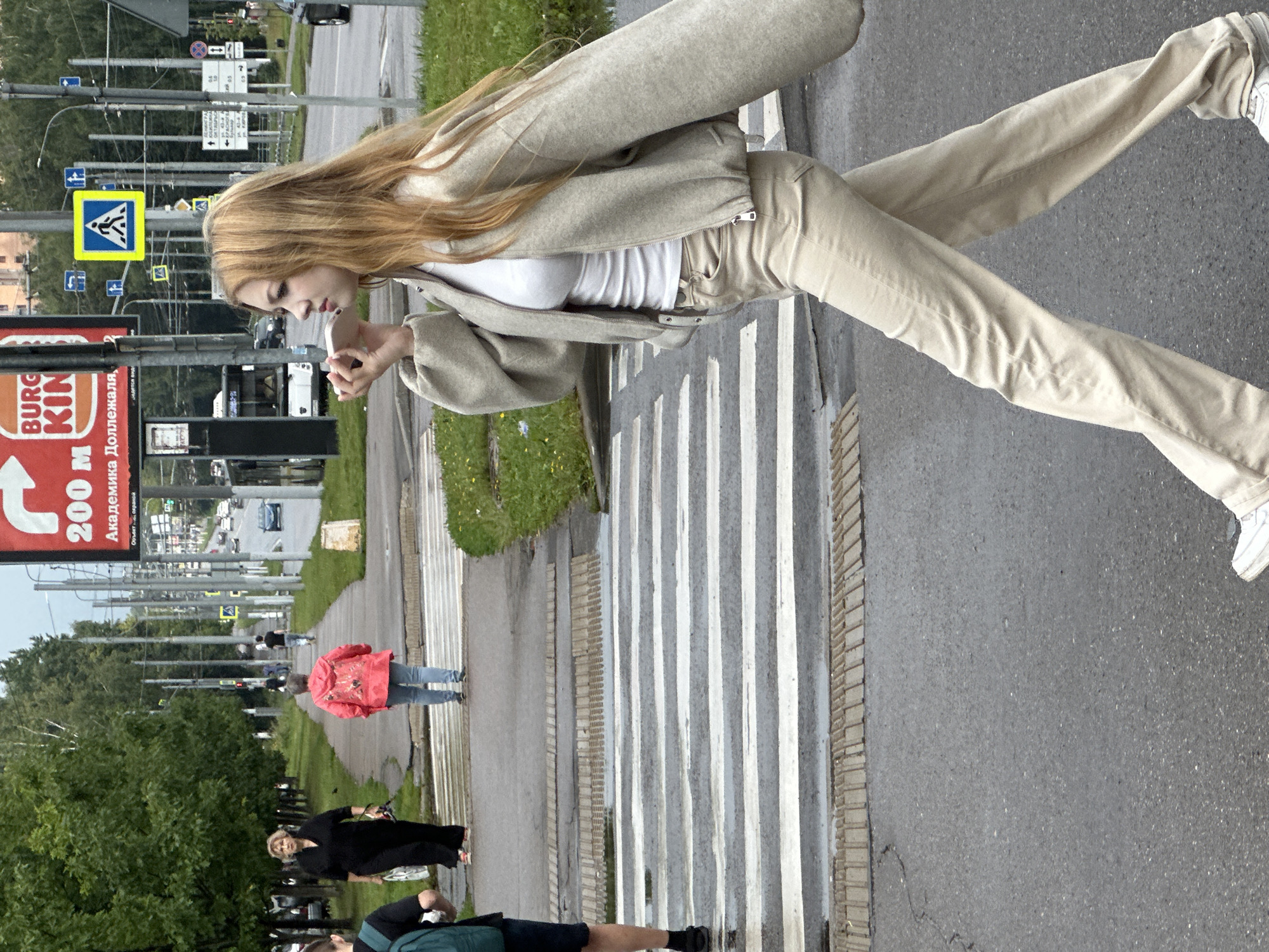 This is a medium shot of a young woman with long, blonde hair walking across a crosswalk. She is wearing a light-colored bomber jacket over a white t-shirt and tan pants. She is holding a light pink object, possibly a phone, to her ear. Behind her, an older woman in a black dress is walking on the sidewalk. Further back, a person in a red jacket is also walking on the sidewalk. The crosswalk is wet, suggesting recent rain. In the background, there are trees, streetlights, signs, and cars on a road. One sign visible is for Burger King. This appears to be a street scene in Podolsk, Russia, indicated by the presence of Russian text on signs.