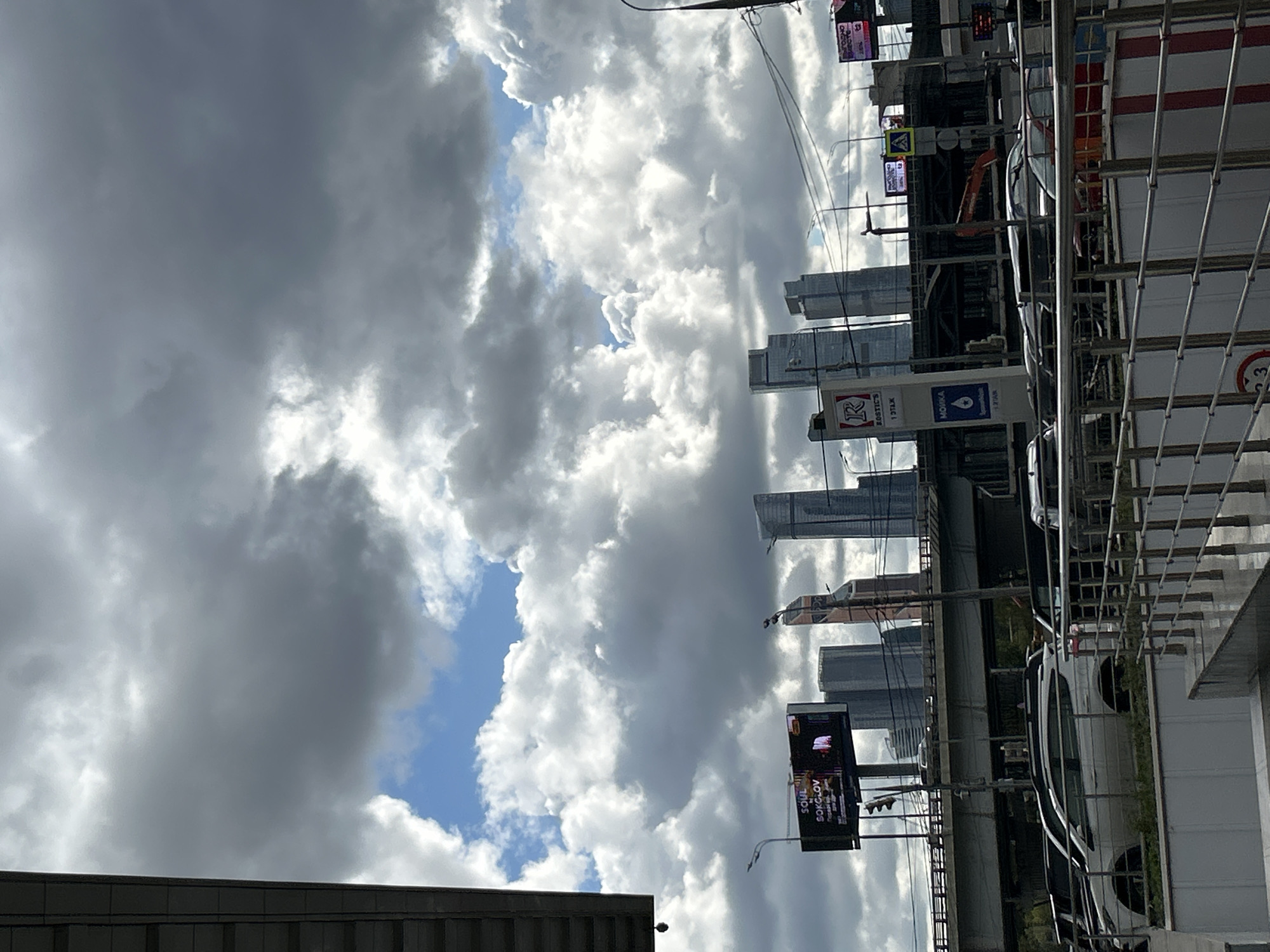 The image shows a cloudy sky over a city scene in Khimki, Russia. In the foreground, there is a metal railing. Behind the railing, several cars are parked. In the middle ground, there is an elevated road or bridge. On the other side of the bridge, there are several tall buildings, some of which appear to be skyscrapers. One building has a large billboard displaying text and an image. There are also streetlights and traffic signals visible. The overall impression is of an urban environment on a cloudy day.